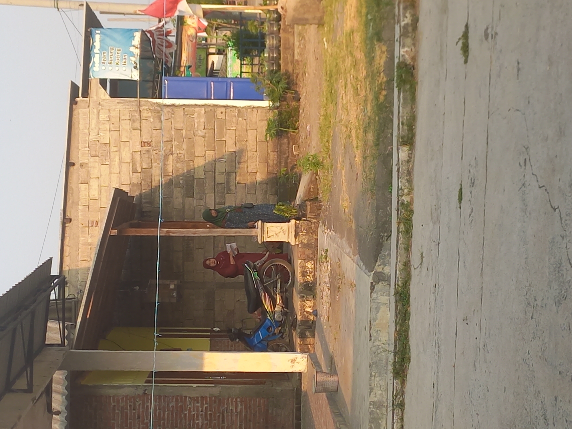 The image depicts a street scene in Pulo Gedang, Indonesia, featuring a brick building and a covered porch area. Two women, both wearing hijabs, are standing near the entrance of the building. One woman, dressed in a maroon hijab and dress, is holding a document. The other woman, wearing a green hijab and a patterned dress, stands to her right. A blue motorcycle with colorful decals is parked on the porch. A grey trash bin is situated to the left of the porch. A blue door is visible on the side of the brick building, and a sign with Indonesian text, listing "Ayam," "Burung," "Kucing," and "Ikan," is hanging from the building. The environment appears to be a residential or commercial area with a paved road in the foreground and some vegetation.