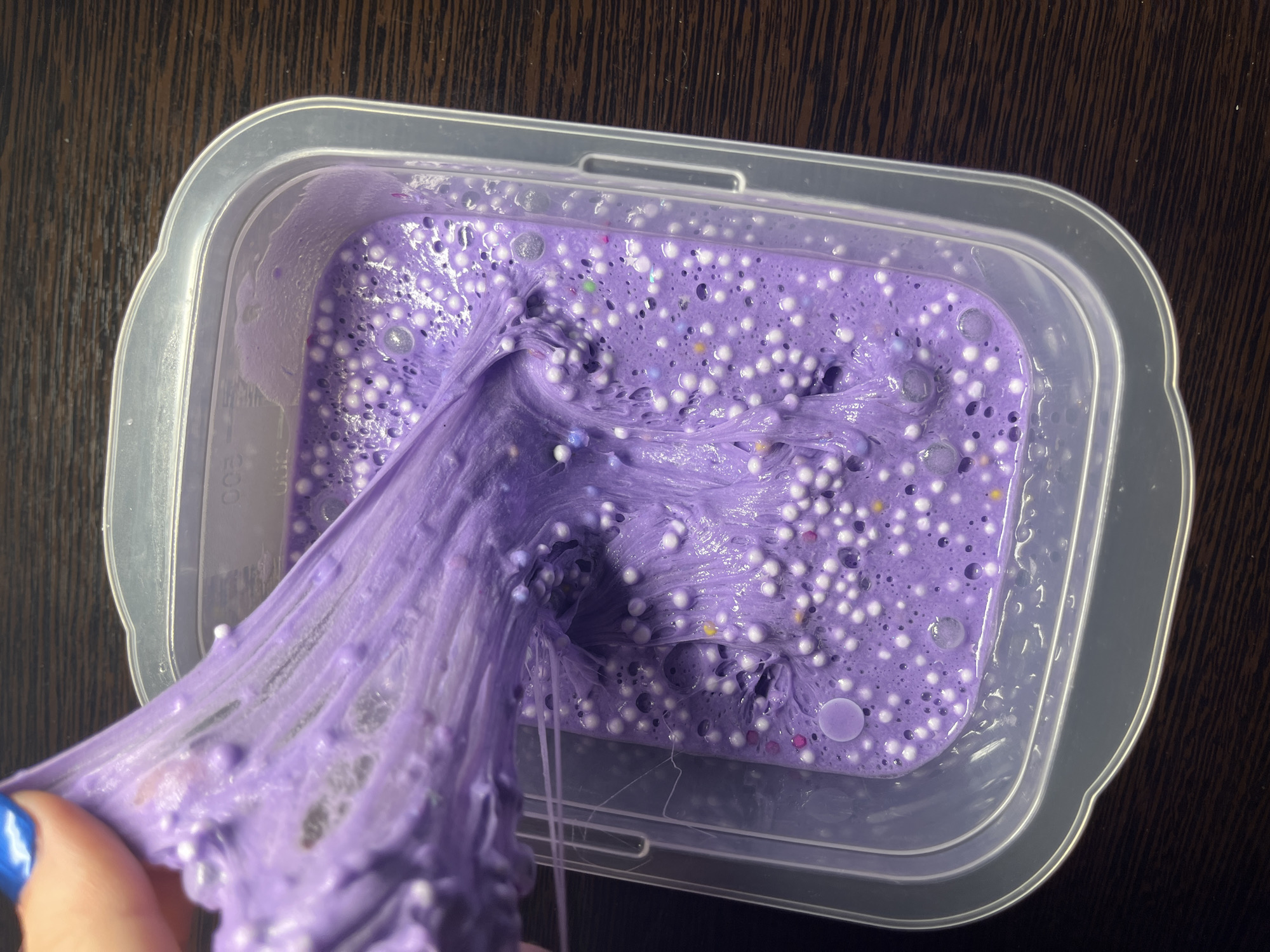 The image depicts a container of purple slime with white beads and small, colorful specks on a dark brown surface. A portion of the slime is being lifted from the container, revealing its stringy texture. The container is clear plastic. The location is not discernable from the provided image.