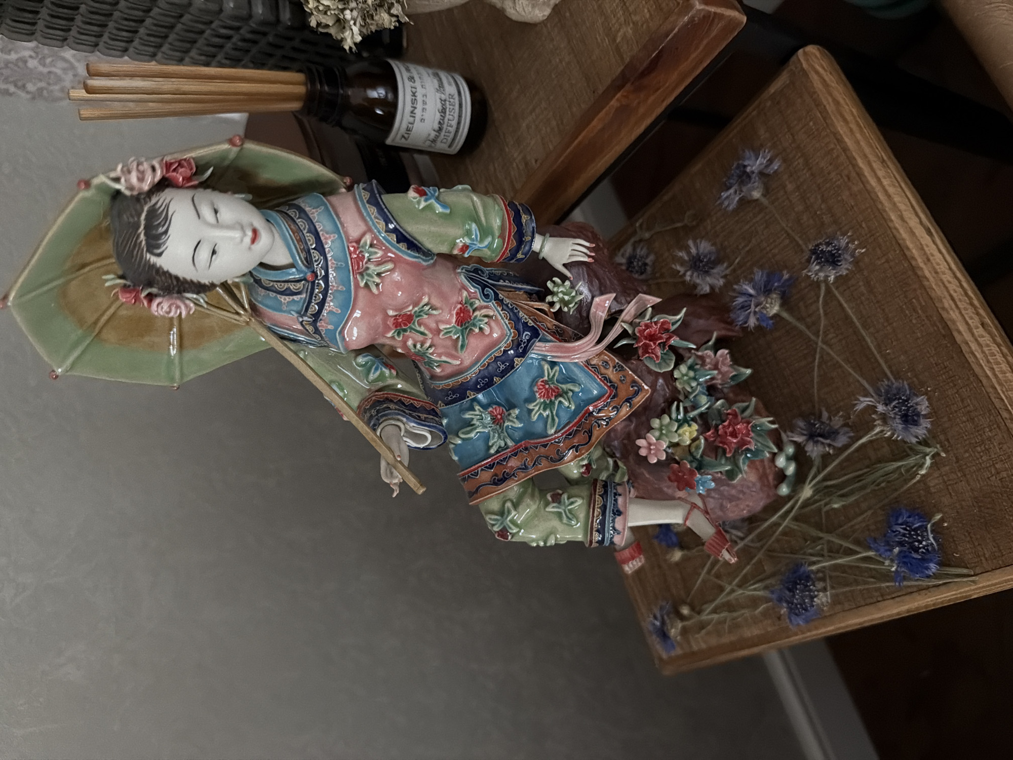 A porcelain figurine of a woman in traditional East Asian attire is displayed on a wooden surface, possibly a table or shelf. She is seated, holding a parasol over her shoulder and is adorned with colorful floral patterns. Dried blue wildflowers are scattered around the base of the figurine and on the wooden surface. To the right of the figurine, a brown glass bottle with a label that reads "ZIELINSKI & SON" and "DIFFUSER" is placed alongside a bundle of wooden reeds, suggesting it is an aroma diffuser. The background appears to be a textured wall.
