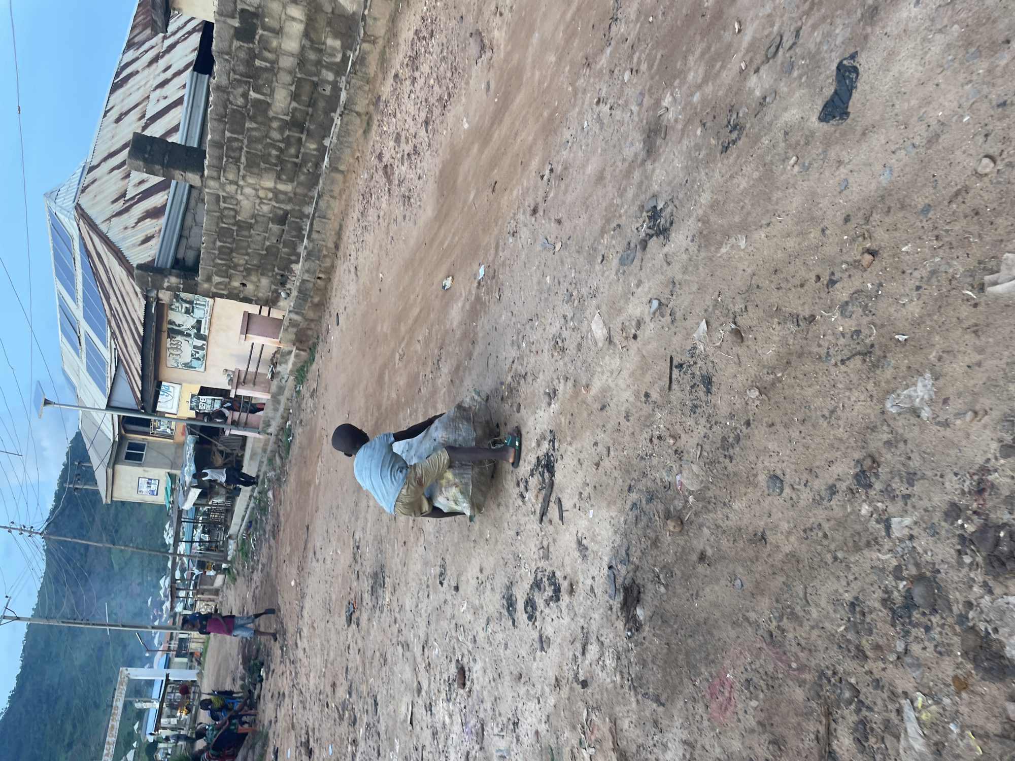 In Lokoja, Nigeria, a young boy, dressed in a light blue t-shirt and beige shorts, is seen from behind, struggling to drag a large, bundled sack of what appears to be refuse across a dusty, unpaved street. The sack is positioned against his legs and he is leaning forward, exerting effort to move it. He is wearing a pair of green and black flip-flops. The street is littered with debris, including small stones, dirt, and scattered pieces of trash. In the background, several buildings line the street, some with corrugated metal roofs, and one structure has solar panels installed. A few people are visible in the distance, including a group seated on the left side of the frame and individuals walking or standing near the buildings. The environment suggests a semi-urban or rural setting, with a backdrop of a hill or mountain. The overall scene depicts a task of waste management being carried out by a child in a modest environment.