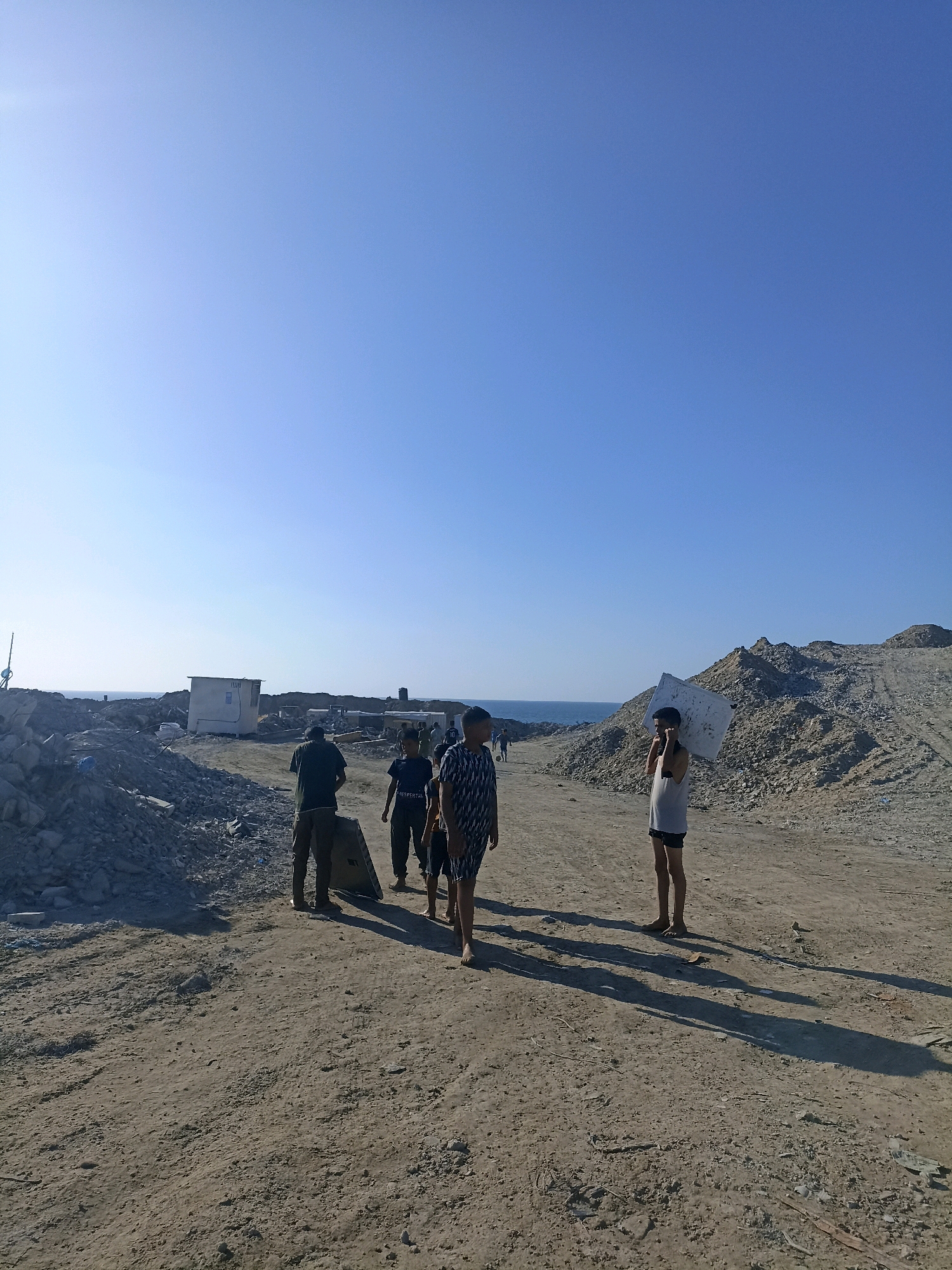 In the city of Gaza, Palestinian Territories, a group of individuals are seen walking on a dusty, dirt path in a desolate, debris-strewn area. The sky is clear and blue. In the background, mounds of rubble and construction materials suggest destruction or ongoing reconstruction. A small, white portable building with a dark door and window is visible on the left side of the frame, near a pile of grey rocks. Further in the distance, the sea can be seen under the bright sky.
There are at least five people visible. In the foreground, to the right, a young boy wearing a white tank top and dark shorts is carrying a large, flat, white object over his shoulders. To his left, two other individuals are walking away from the camera. One appears to be a young person in a patterned black and white shirt and dark shorts. Another, slightly behind and to the left, is wearing a dark t-shirt with white lettering that reads "RESPETAR." Further to the left, a person in a dark green shirt and dark pants is facing away from the camera. A fifth person, partially obscured, is walking behind the person in the dark green shirt. The ground is uneven and covered in dirt, small stones, and scattered debris. The overall atmosphere of the scene is one of hardship and a rugged environment, likely a result of conflict or environmental challenges.
