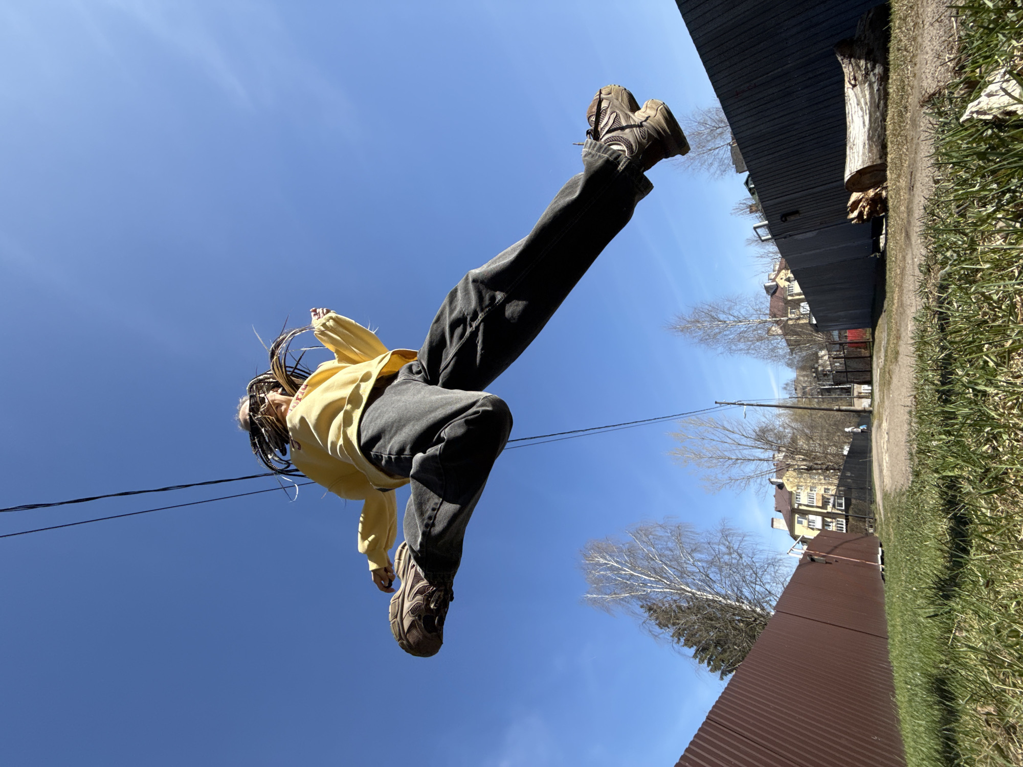 The image captures a person in mid-air, dynamically positioned horizontally across a bright blue sky. The individual wears a yellow long-sleeved top, dark gray or black pants, and brown athletic shoes. Their hair appears to be in braids or dreadlocks.
The primary background is a clear, bright blue sky, with minimal, wispy white clouds. A black utility wire stretches across the upper left part of the frame. On the right side, a dark corrugated metal fence runs vertically, alongside a dirt path where a large wooden log rests. Green grass is visible in the bottom right foreground. Beyond the fence, several leafless trees and distant multi-story residential buildings with multiple windows are visible under the sunny conditions.
The photograph is taken from a low angle, looking upwards at the person against the sky, and is rotated 90 degrees clockwise, giving the impression of horizontal flight. If correctly oriented, the person would be seen jumping upwards. The scene takes place during a sunny day, likely in spring given the bare trees. The setting suggests a residential or semi-rural area, consistent with the location context of Bor, Russia. There is no visible text in the image.