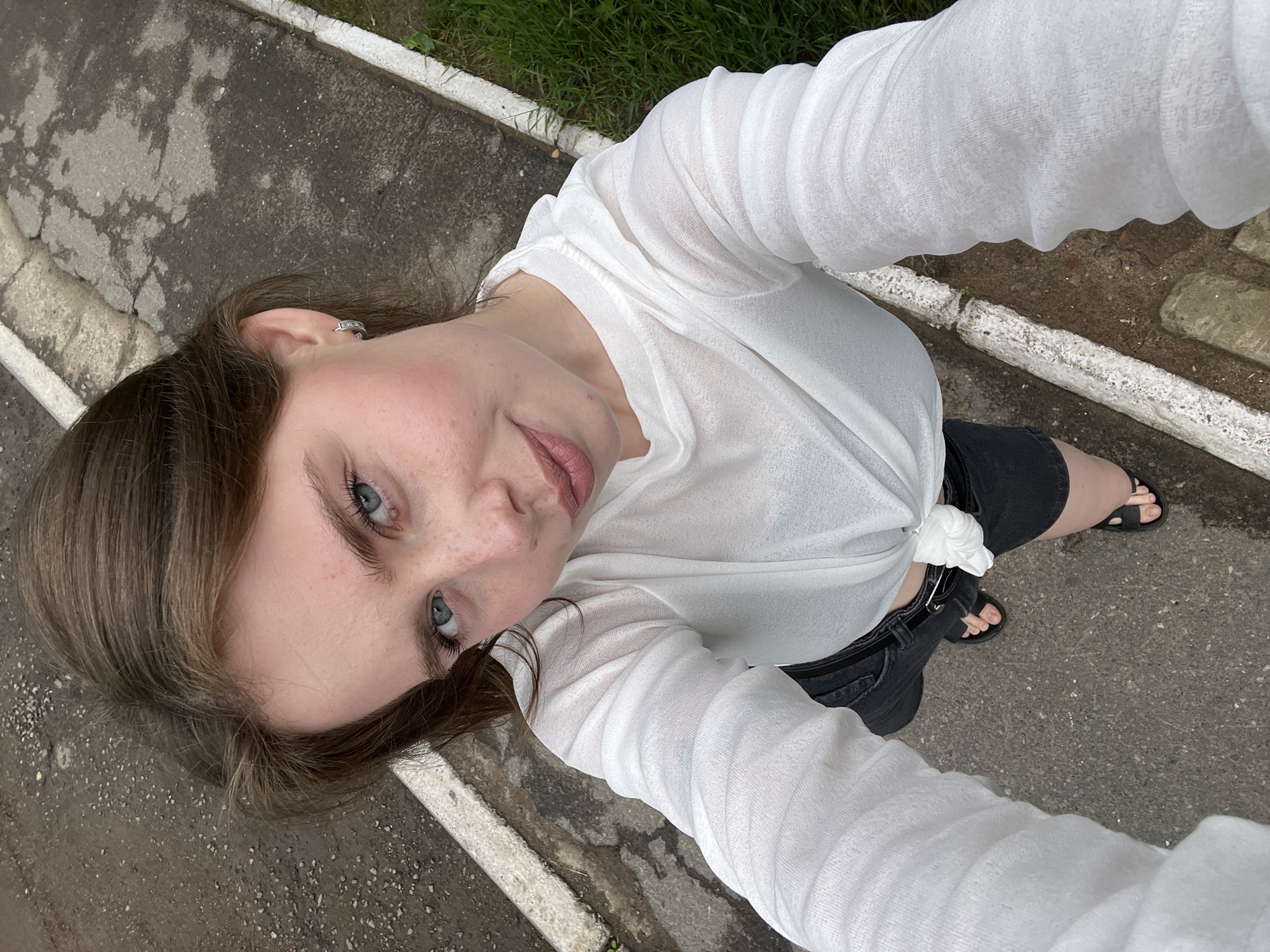 A young woman with light brown hair and blue eyes is taking a selfie outdoors. She is wearing a white, sheer long-sleeved shirt tied at the waist and black shorts. Her face is turned towards the camera, with a slight smile. She is wearing small hoop earrings. The background shows a cracked asphalt surface with a white curb and green grass to the right. The location appears to be a street or pathway in the city of Chaikovsky, Russia.