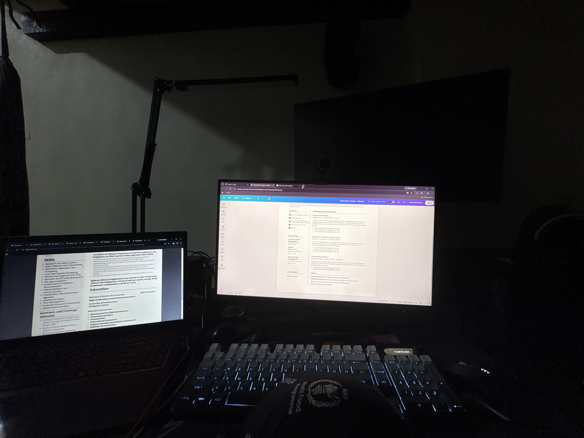 This is a dark, indoor scene featuring a desk setup with two computer monitors, a laptop, and a mechanical keyboard. The main monitor displays a resume, likely being edited, with sections for skills, education, and professional experience. A desk lamp illuminates the scene from the left. The laptop to the left also shows a portion of the resume. The overall environment appears to be a home office or study area. Based on the resume content visible on the screen, specifically the mention of "The Effects of Typhoon Frequency on Food Price Inflation in the Philippines: An AREL-based modeling Approach," and the presence of local curriculum vitae formatting, the location is likely Davao City, Philippines, given the common use of such academic topics in research and documentation within the country. No individuals are visible in the image.
