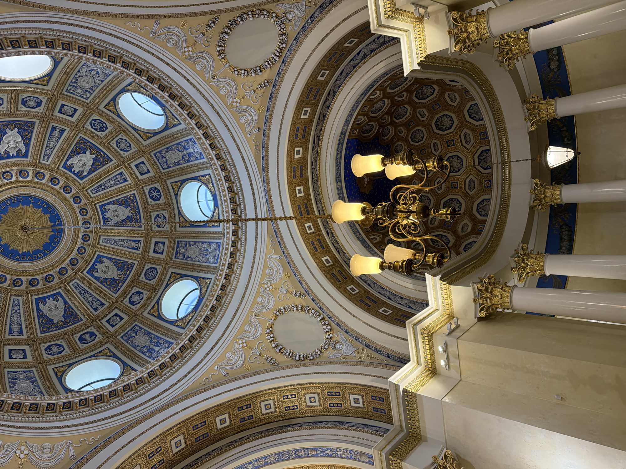 The image shows the interior of a grand dome, likely in a public building such as a museum, government building, or religious site, located in посёлок дачного хозяйства «Архангельское», Russia. The dome is elaborately decorated with intricate patterns and motifs in gold and blue. Oval windows are visible in the upper part of the dome, allowing natural light to filter in. A multi-tiered chandelier hangs from the center of the dome, illuminating the space with warm light from its globes. The lower parts of the image reveal white columns with ornate gold capitals, supporting the structure. The overall impression is one of opulence and historical significance.