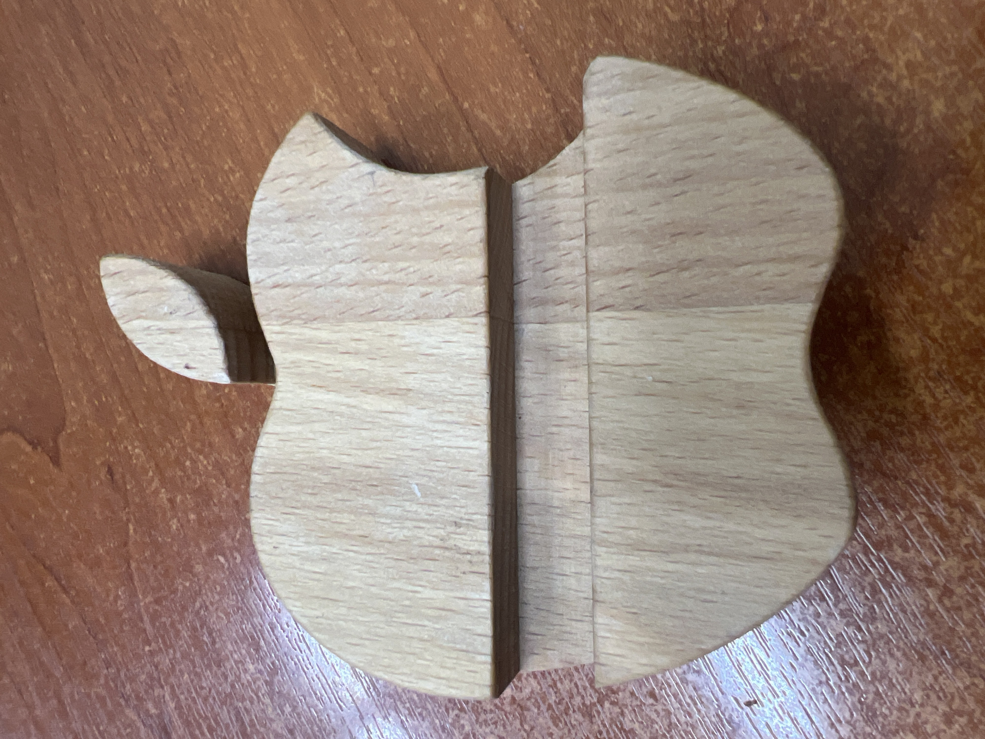 A wooden, unfinished-looking, shield-shaped object rests on a wood-grain surface. The shield shape has an additional piece to the left, resembling a wing. The main part of the shield is divided into two halves separated by a vertical groove. The location in Chaikovsky, Russia is unknown as no identifiable location is visible.