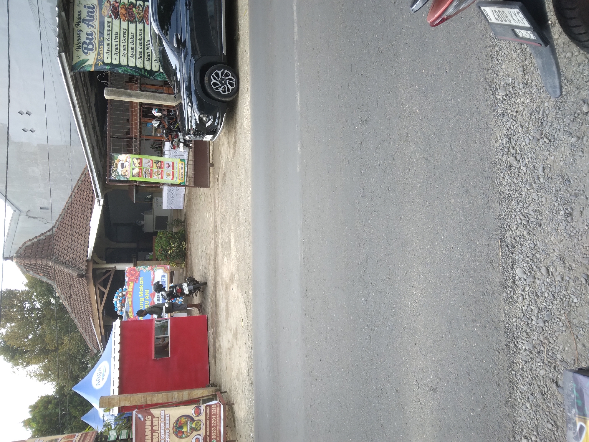 The image depicts a street scene in Desa Pamijen, Kecamatan Sokaraja, Indonesia, likely in front of a small eatery. A black SUV is parked on the right side of the frame, its front fender and wheel clearly visible. To the left of the SUV, a dark-colored motorcycle is parked. A man, wearing a dark shirt and shorts, stands near the motorcycle, facing away from the camera. He appears to be interacting with a small red structure with a window, possibly a food stall or kiosk, which has a blue tarp canopy above it.
Behind the red structure and the man, a building with a pitched, tiled roof is visible. To the right of this building, a larger structure with a metal roof extends, serving as a covered area for the eatery. A banner advertising "Warung Makan Bu Ani" (Bu Ani's Eatery) is prominently displayed, listing various chicken dishes and fish as offerings. Another banner, partially obscured by the red kiosk, also advertises food items and a phone number.
The environment appears to be a roadside setting. The ground in front of the eatery is a mix of gravel and dirt. The road itself, a paved surface, occupies the lower half of the image. The overall impression is that of a local business operating along a public thoroughfare.