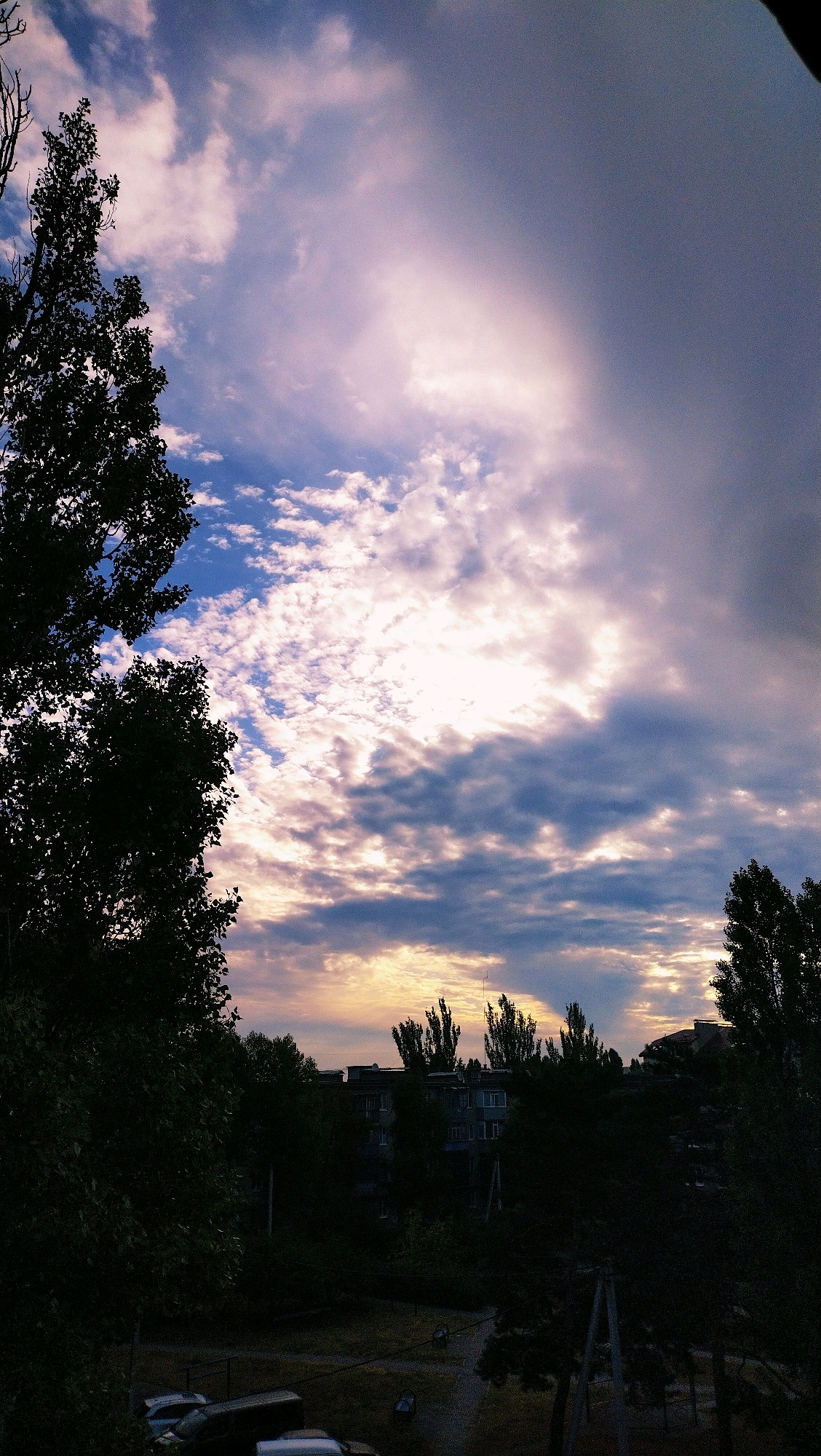The image depicts a dusk scene in Horishni Plavni, Ukraine. The sky is a dramatic mix of dark clouds and patches of pale pink and yellow light, suggesting the sun has recently set or is about to rise. Silhouetted against this sky are the dark forms of trees, some with dense foliage and others bare branches, framing the view. In the midground, several multi-story buildings, typical of Soviet-era apartment blocks, are visible, also largely in silhouette. A few scattered lights can be seen in the windows of these buildings. The foreground is dark, with the shapes of parked vehicles, including cars and vans, barely discernible. The overall impression is one of a quiet urban evening.