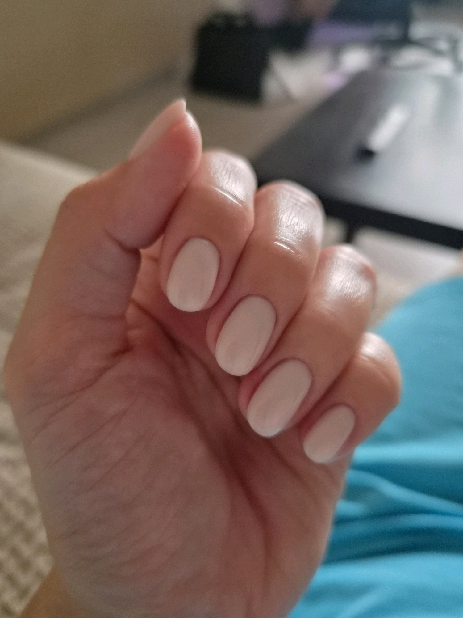 The image shows a close-up of a person's left hand with light beige-colored fingernails. The hand is positioned in the foreground, with the fingers slightly curled. The background is out of focus and appears to be an indoor setting. Part of a couch or upholstered furniture is visible on the left, and a dark surface, possibly a table, is in the background on the right. A bright blue fabric is visible on the lower right side of the image. There is no visible indication of the city of Kudrovo, Russia, or any specific location details. The lighting suggests it may be taken indoors, possibly during the day.