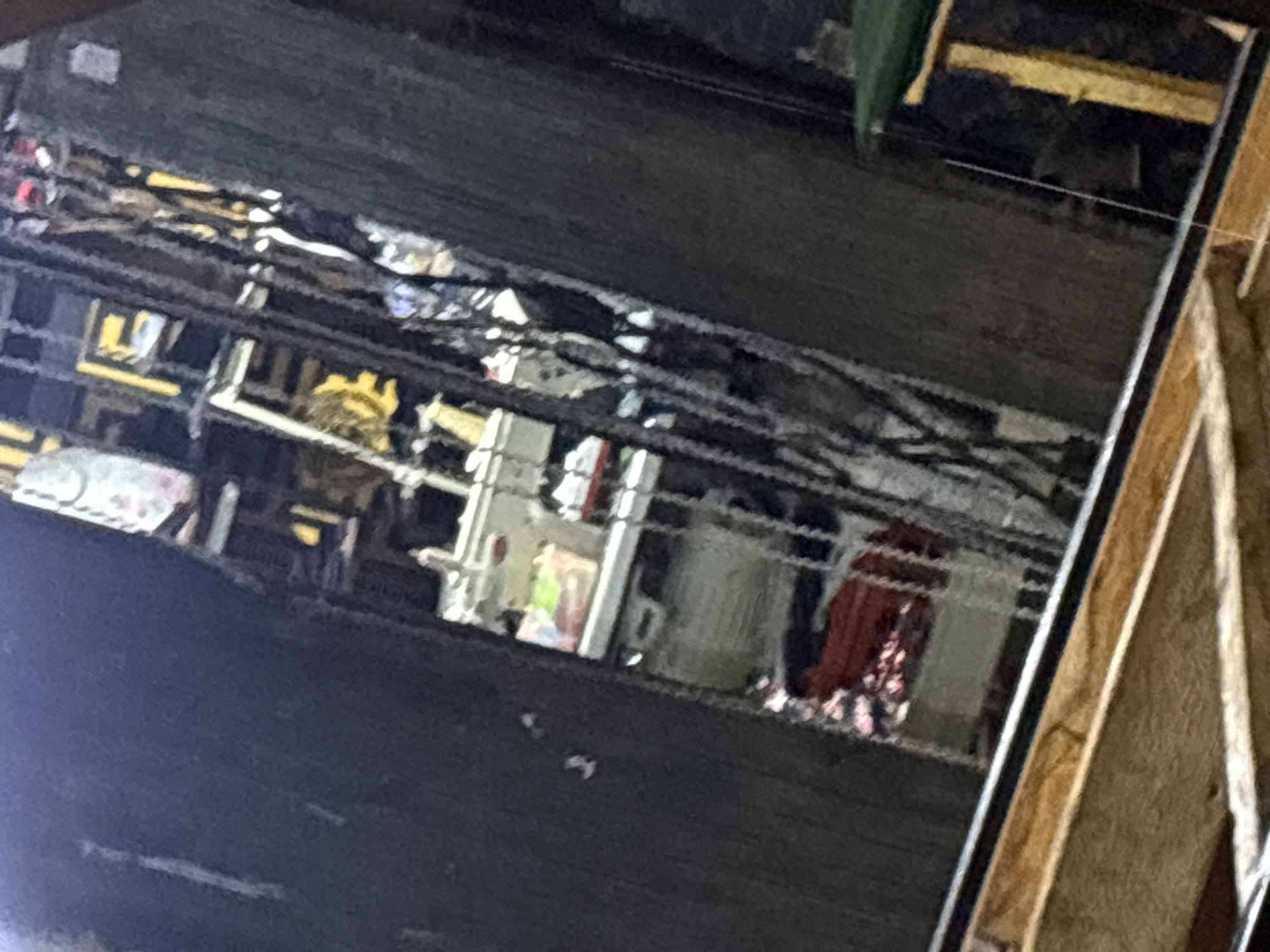 The image shows a reflection on a dark, possibly lacquered, surface, likely a tabletop or similar. The reflection reveals an interior space with various objects. Visible are furniture pieces, including what appears to be shelving or storage units, and a table covered with a patterned cloth. Other indistinct objects, possibly household items or clutter, are scattered throughout the reflected space. The overall lighting in the reflected scene is somewhat dim. To the right of the frame, a wooden structure is visible, suggesting construction or framing work. The location (Bukuru, Nigeria) is not discernable from the image.