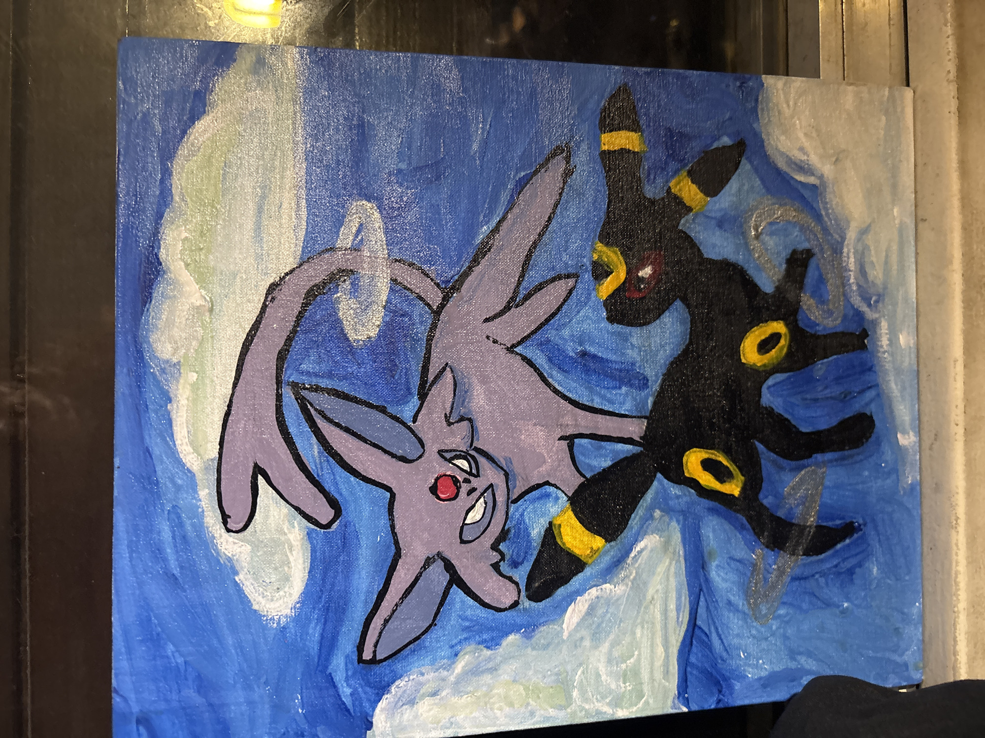 A hand-painted artwork on canvas depicts two stylized cartoon-like figures, resembling the Pokémon Espeon and Umbreon, against a swirling blue and white background. The purple figure, Espeon, is positioned on the left, largely inverted, with a red gem on its forehead, a slight smile, and a long, forked tail with a white ring. The black figure, Umbreon, is on the right, featuring red eyes and prominent glowing yellow rings on its forehead, ears, and legs.
The two figures are dynamically intertwined, suggesting interaction or a spirited pose within an abstract, ethereal environment. The background is composed of various shades of blue, ranging from dark to medium, blended with soft white and subtle light green cloud-like formations, possibly representing a mystical sky or an energy field. Visible brushstrokes indicate the artwork is likely executed with acrylic or oil paints. The color palette is vibrant, utilizing strong contrasts such as the purple and black characters against the blue background, accented by bright yellow and red details. A stylized silver mark, possibly a signature, is visible on the lower right area of the painting, near the Umbreon figure. The overall impression is one of dynamic fantasy art. This artwork is located in Buenos Aires, Argentina.