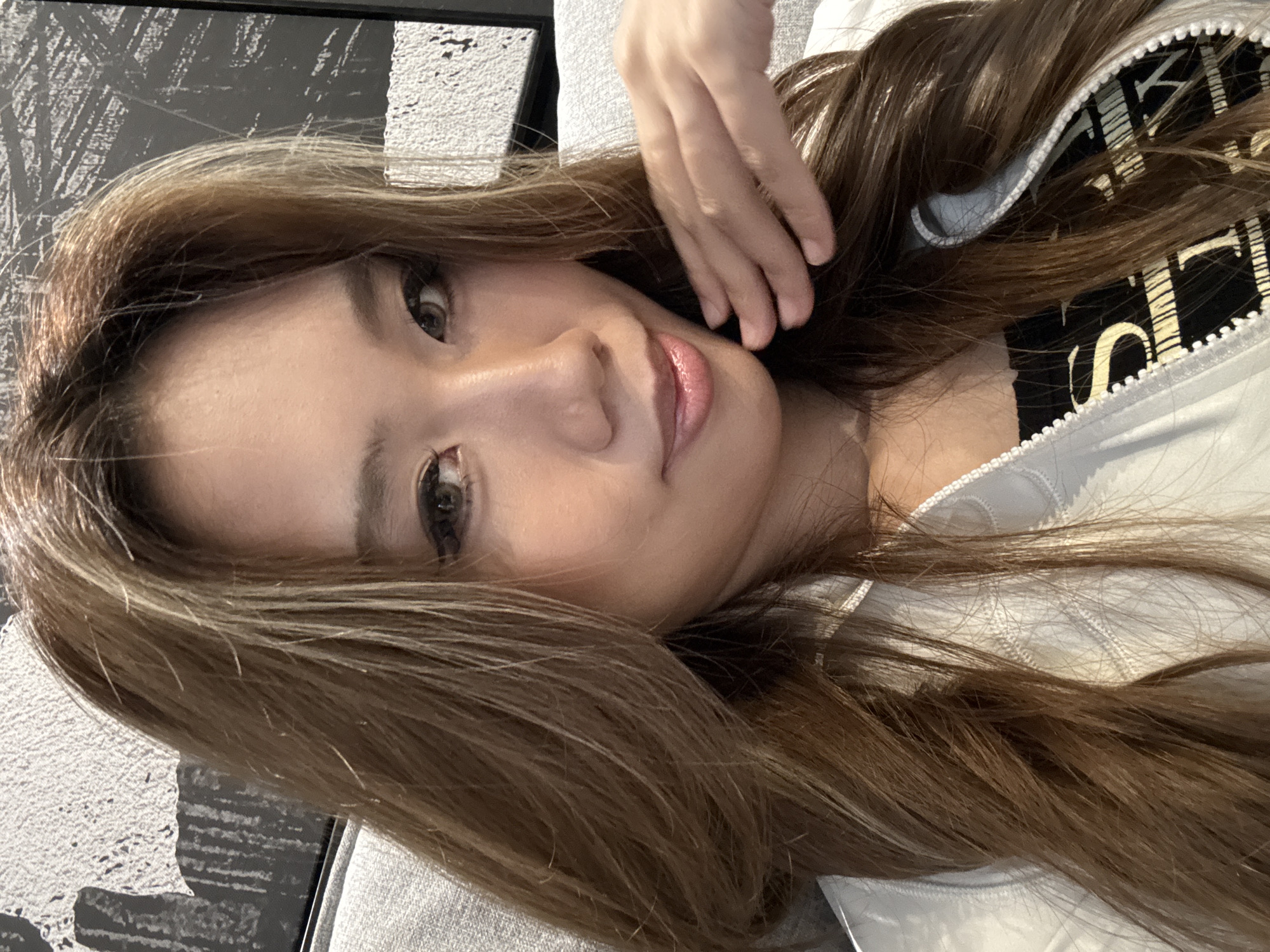 A woman lies on a gray surface, possibly a couch or bed. She has long, brown hair with blonde highlights, and fair skin. Her right hand is near her face. She wears a light-colored jacket, partially unzipped, revealing a black shirt with a light-colored design. The background includes a black-and-white patterned wall or artwork. There's no information in the image that definitively places it in Liangjiang, China.