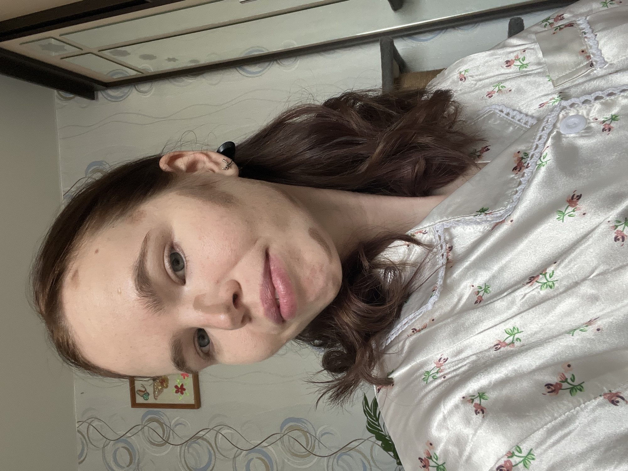 A medium shot, taken indoors and possibly in a bedroom, shows a woman with fair skin, light eyes, and brown hair. Her hair is partially tied back, revealing an earring. She is wearing a satin pajama top with a floral pattern and white lace trim. In the background, there is patterned wallpaper with circular designs. A framed picture hangs on the wall to the left of the woman's face, close to the bottom of the frame. The top of a cabinet or closet is visible at the top of the frame, suggesting that the photo was taken at a slight upward angle. Given the user's request, and as the user resides in Chaikovsky, Russia, it can be inferred that the photo was taken in that location.