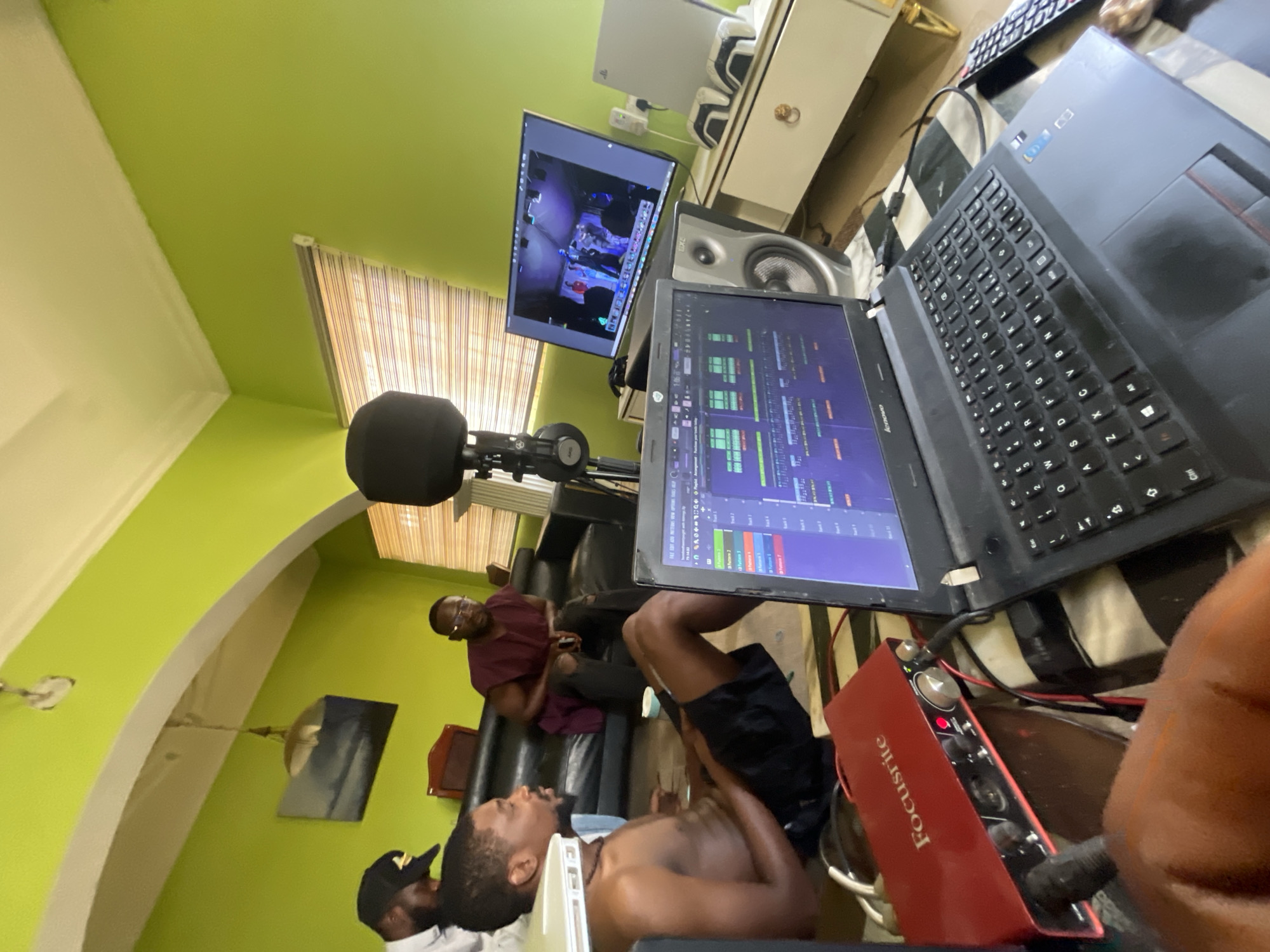A horizontal, eye-level shot shows a man lying on a couch in a studio-like setting, appearing shirtless, and in a room with green walls and cream-colored trim near a partially visible ceiling. Another man sits upright behind him on the couch. In the foreground, a laptop displays audio editing software, and a red Focusrite audio interface is visible. Above the laptop, a monitor displays video footage. A microphone and pop filter are positioned to the left of the monitors. The room appears to have blinds covering the window, and a white dresser and other equipment are present. The image was likely taken in Abuja, Nigeria, based on the details provided.