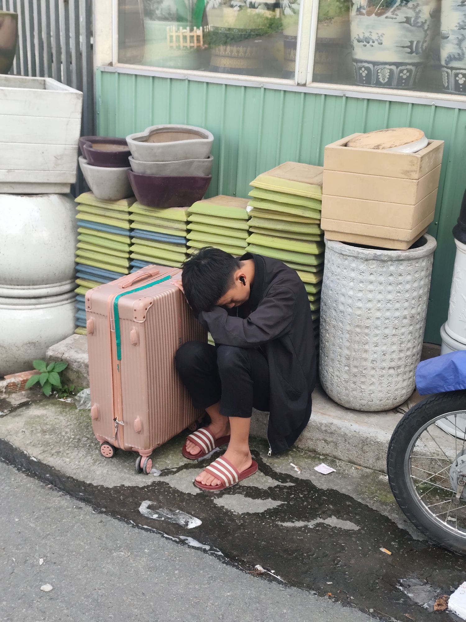A young person wearing a black shirt and pants, with earbuds in, is sleeping while leaning against a light pink suitcase. They are wearing flip-flops with red and white stripes. The suitcase is positioned on a paved surface next to a concrete curb. Behind the suitcase, stacks of green and yellow square-shaped items, possibly plant pot saucers or trays, are piled high. To the left of the suitcase, there are large white planters and other decorative pots. To the right of the suitcase, a large, textured white pot stands prominently, and further to the right, a motorcycle wheel is partially visible. The scene appears to be outdoors, likely in a marketplace or a storage area for garden supplies, given the abundance of planters and gardening accessories. The overall impression is one of a traveler resting during the day. The lighting suggests daytime. The location is specified as Phường Xuân Hương - Đà Lạt, Vietnam. There is no visible text in the image.