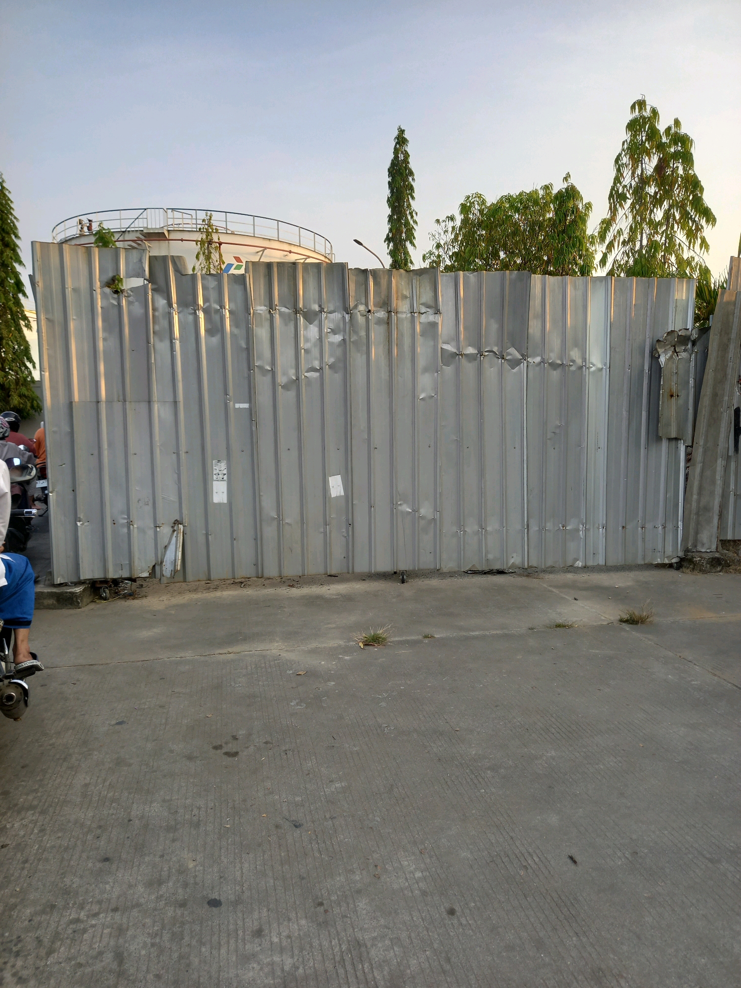 The image depicts a large, corrugated metal fence in a city setting, likely in Samarinda, Indonesia. The fence appears weathered and dented, with some sections showing damage. Behind the fence, a portion of a large, white cylindrical storage tank with a railing around its top is visible. The tank has some markings, including what appears to be a red and blue logo. Several trees with green foliage are present behind the fence, with some taller, slender trees on the left and a cluster of wider trees on the right. The sky is a pale blue with hints of sunset colors. In the foreground, a concrete pavement is visible, with some cracks and scattered patches of dry grass. On the left side of the frame, a person is seated on a motorcycle, wearing blue pants and white footwear. Another motorcycle with a rider wearing an orange shirt and a black helmet is partially visible further down the road. The overall scene suggests an industrial or storage area on the outskirts of a city, possibly near a port or fuel depot.