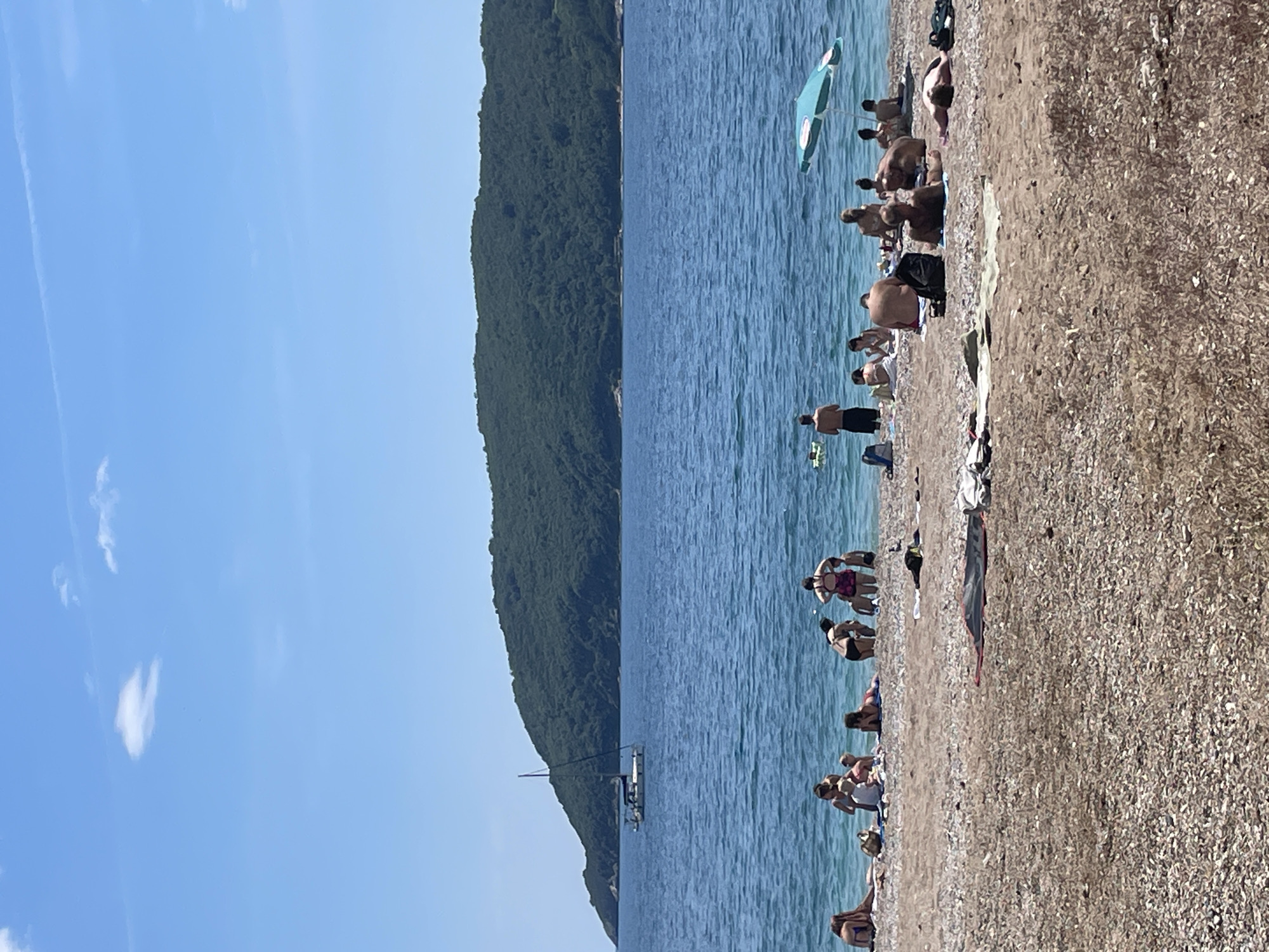 The image shows a beach scene in Budva, Montenegro, densely populated with people enjoying a sunny day. The beach is composed of small pebbles and extends along a shoreline with a backdrop of a forested hillside that rises from the sea. The blue sea has a boat in the distance. People are sunbathing, swimming, and generally relaxing. There is a beach umbrella providing shade to a group of people.
