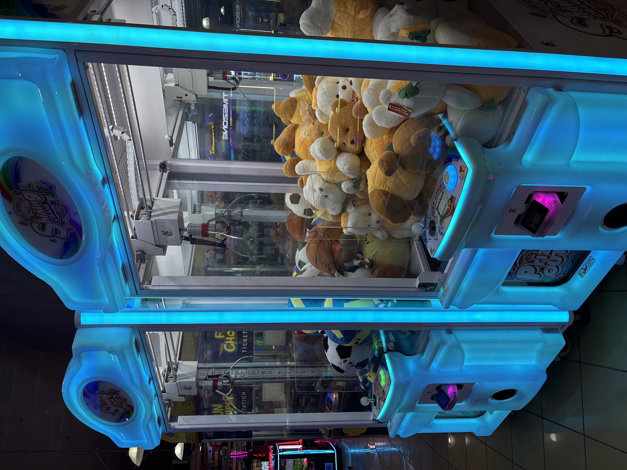 The image shows two brightly lit arcade claw machines filled with plush toys. The machines are located in a brightly lit area, possibly a mall or entertainment center, with other arcade games visible in the background. The claw machines have illuminated blue accents and control panels with joysticks and buttons. The plush toys inside include various animals, such as bears, dogs, and balls. There are no people visible in the image. The location is identified as being in Davao City, Philippines.