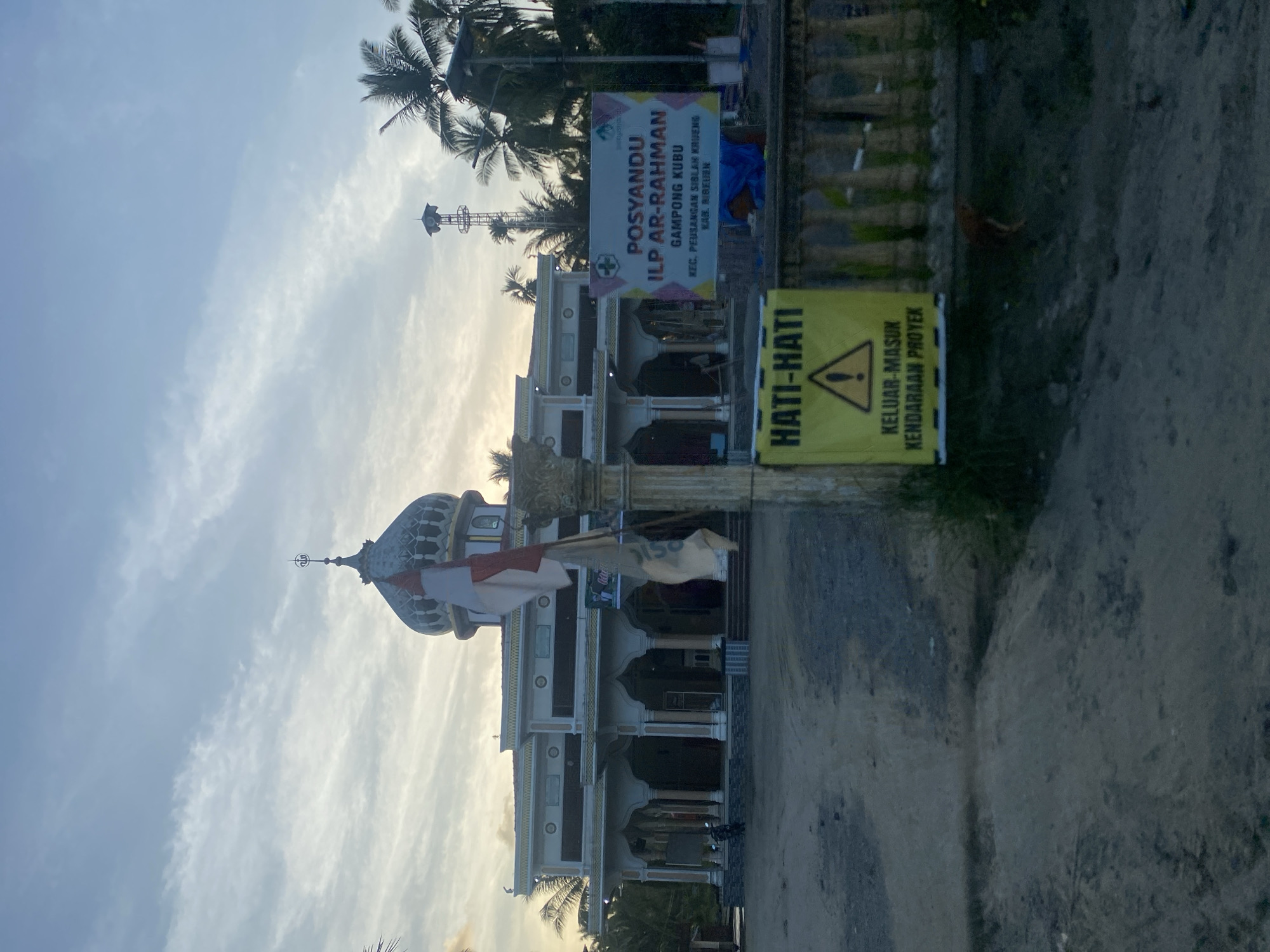 The image presents a mosque-like structure in Kubu, Indonesia, identifiable by the dome and minaret, with a flag fluttering beside it. The building is a light color with a two-story design. A sign reading "POSYANDU ILP AR-RAHMAN GAMPONG KUBU" is visible, indicating a local health service post. A yellow warning sign is present, with the text "HATI-HATI KELUAR-MASUK KENDARAAN PROYEK," alerting passersby to construction traffic. Palm trees are visible in the background against a partly cloudy sky. The scene is situated on a dirt road.