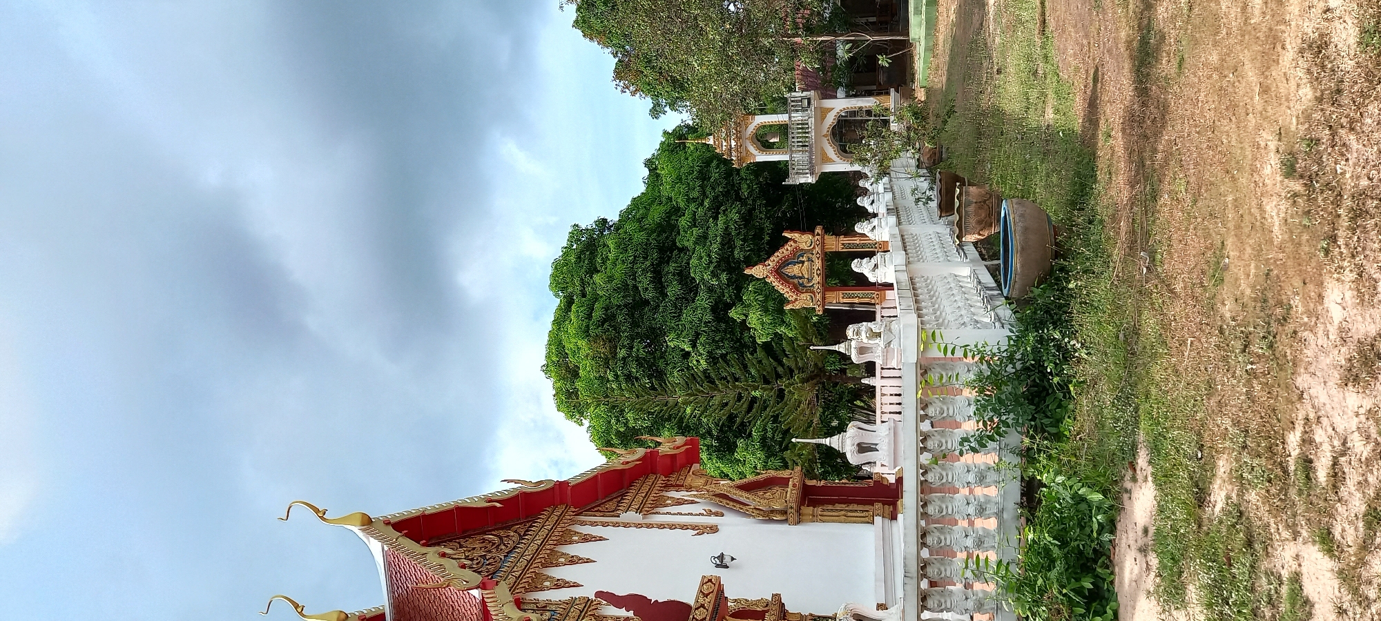 The image shows a religious building in Moo 4, Thailand, likely a Buddhist temple, judging from the architecture. The structure features a red and gold roof, white walls, and intricate decorative patterns. A white balustrade with sculpted figures runs along the side of the building. Behind the structure, there is a large tree and other vegetation. The sky is overcast. Adjacent to the building, a patch of dry, grassy land can be seen. A large, concrete drum with a blue rim is visible near the balustrade.