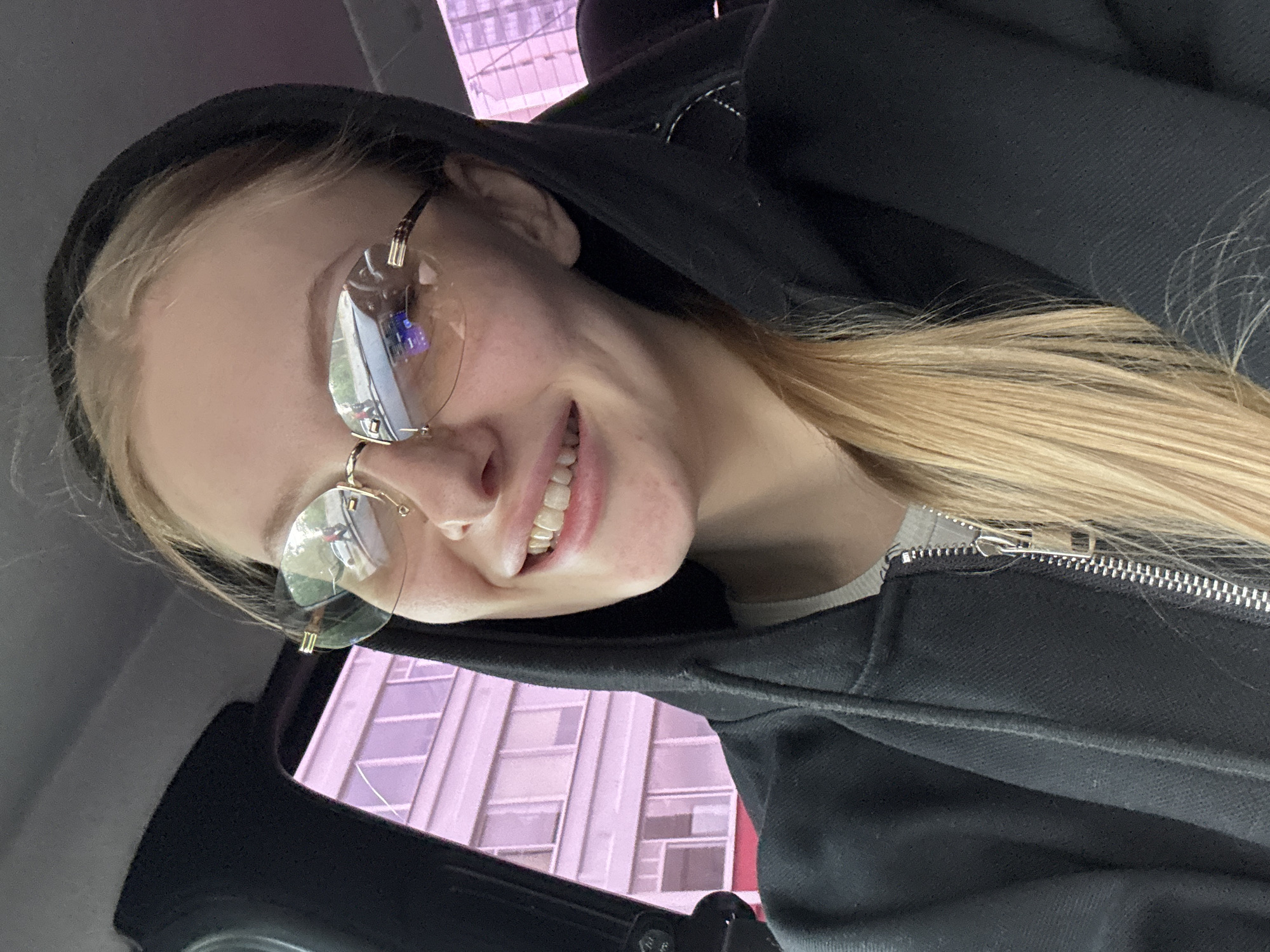 A person wearing a black hooded sweatshirt and rimless glasses smiles towards the camera. Outside the car window, a tall building with repeating rectangular windows is visible. This is Perm, Russia. The person is likely seated in a car, based on the interior car frame around the window. The hoodie zipper is slightly unzipped to reveal a gray undershirt.