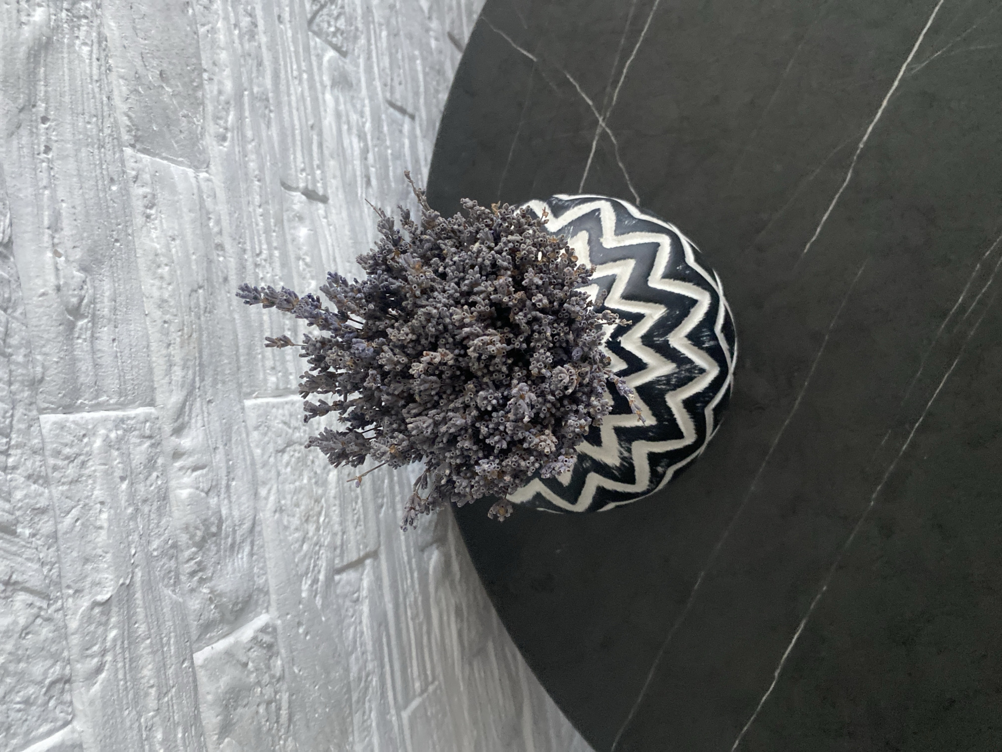 A decorative vase filled with dried lavender sits on a dark, marbled table. The vase has a black and white zigzag pattern. Behind the vase and table is a white brick wall. The image does not provide any information that would allow for determining its location in Chișinău, Moldova.
