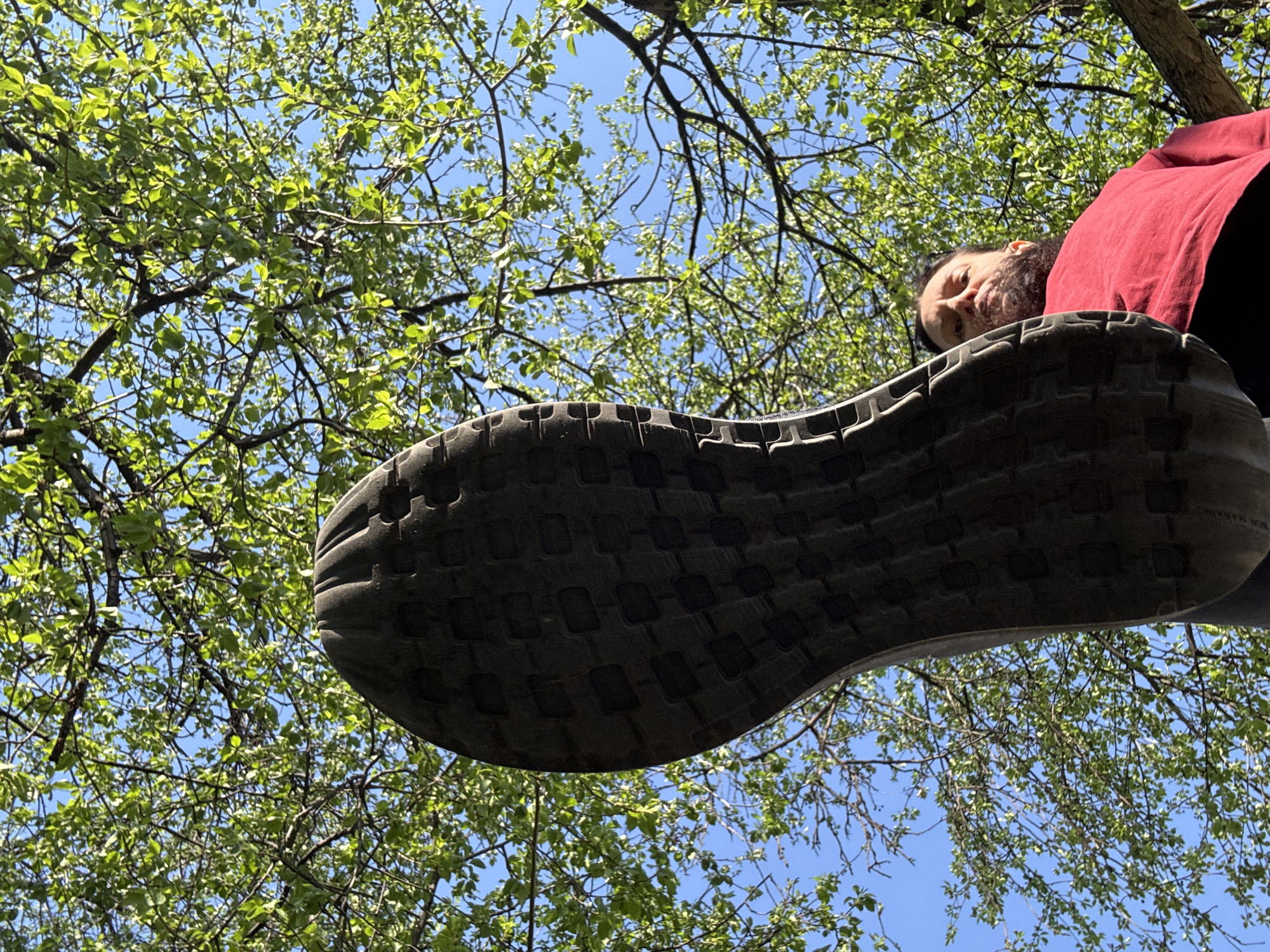 The image is taken from a low-angle perspective looking up at the sole of a shoe and a person. The shoe's sole is the most prominent object, appearing large and with a textured pattern. Behind the shoe and person, the sky is visible through the branches and leaves of a tree. The person's face and part of their torso (wearing a red shirt) are visible. The location in the city of Chaikovsky, Russia is not discernible from the image.