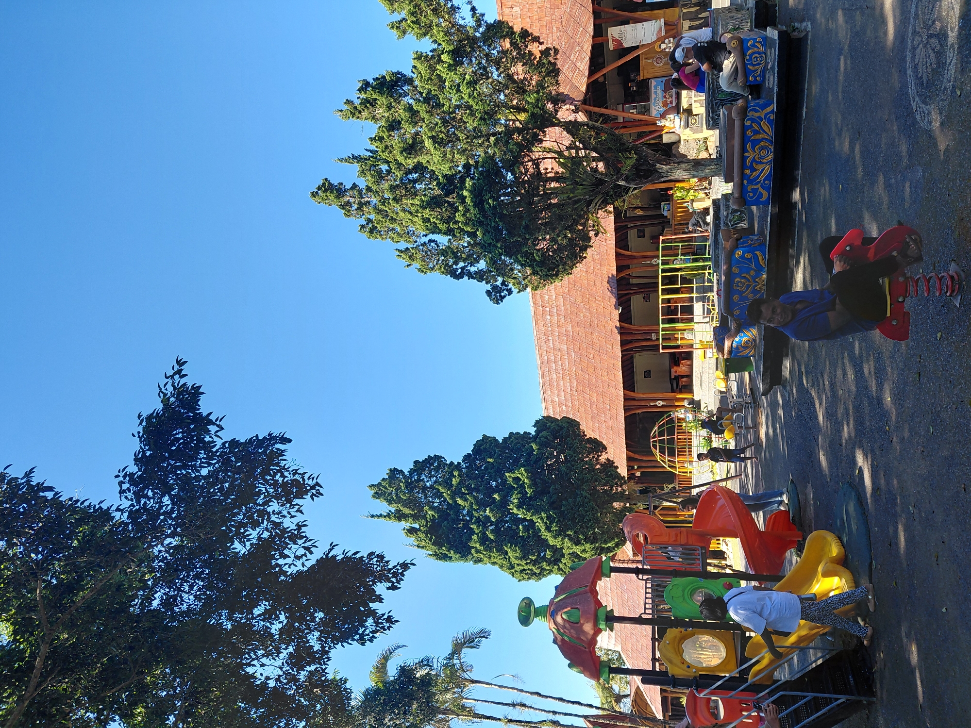 The image depicts a sunny outdoor scene at a playground in Baturiti, Indonesia. The playground features a colorful play structure with slides, a climbing wall, and a bouncy toy. Several people are present, including children playing and adults supervising. In the background, a traditional Indonesian building with a red tiled roof stands, possibly a restaurant or gathering place. Tall trees, including cypress and palm trees, provide shade and add to the tropical ambiance. The ground is paved with asphalt, and scattered leaves indicate it's an outdoor environment.