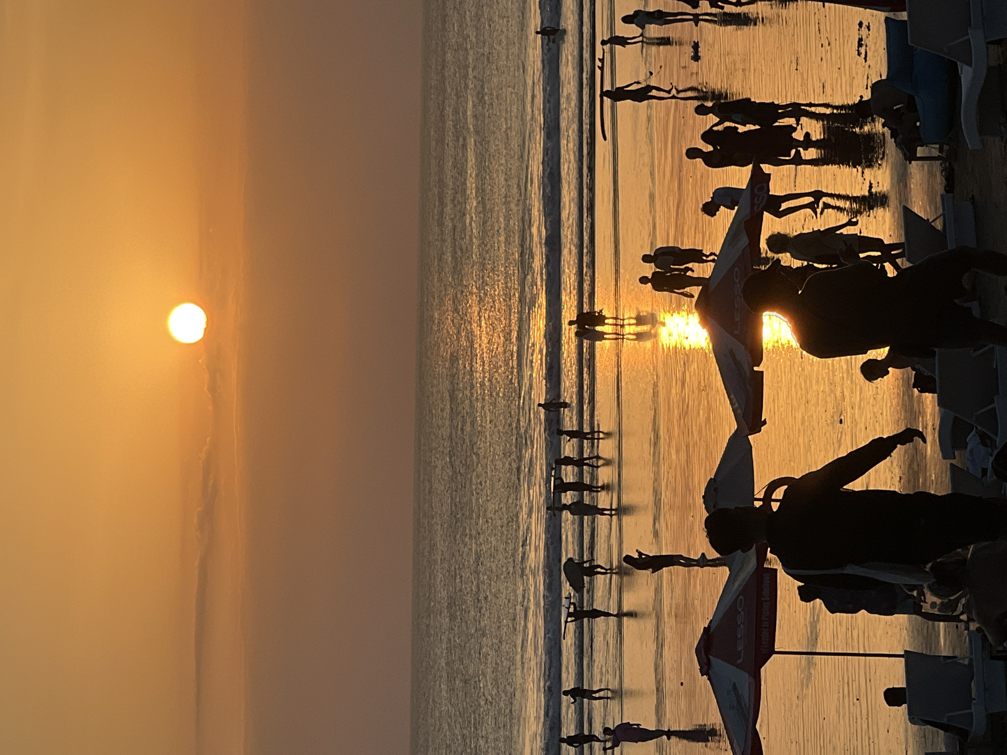 This image depicts a sunset scene at Kuta Beach in Bali, Indonesia. Silhouetted figures of people are scattered across the wet sand and in the shallow water. Some individuals are walking, others are standing, and a few are carrying surfboards. The setting sun casts a warm, orange glow over the entire scene, reflecting brilliantly on the water and the wet sand. Two beach umbrellas, one with the word "LESSO" visible, are present in the foreground. The ocean is calm with gentle waves rolling onto the shore. The overall atmosphere is one of leisure and natural beauty, with the sky transitioning from a bright orange near the sun to a deeper, dusky hue towards the horizon.