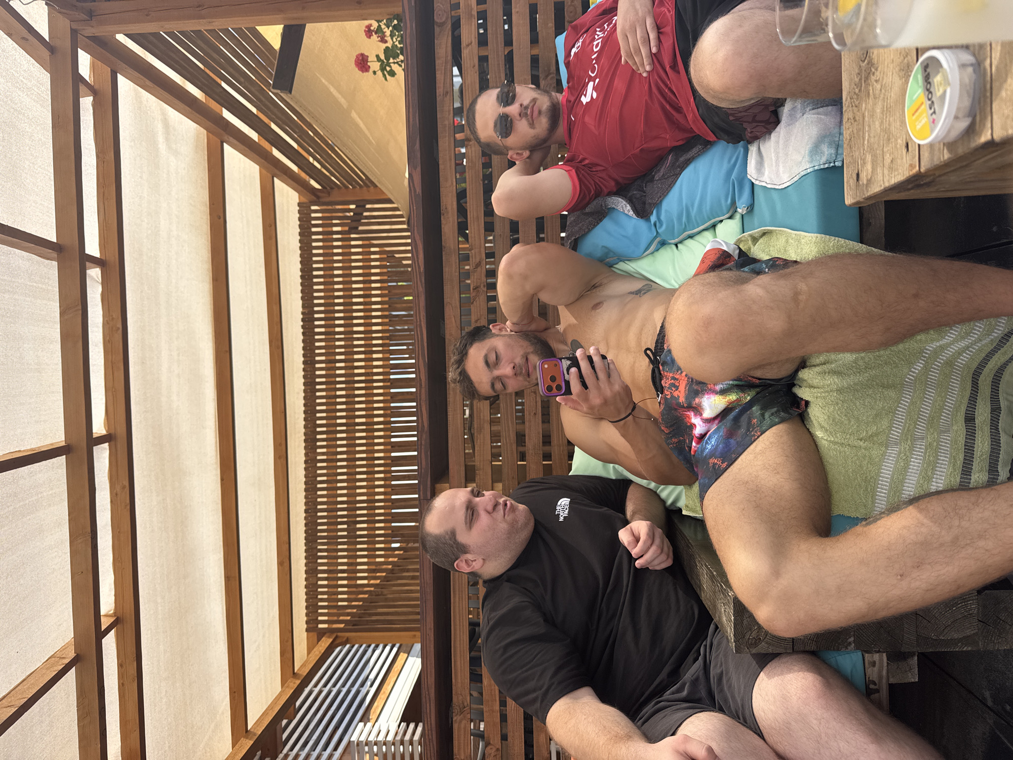 Three men are seated on a blue couch under a wooden pergola with a white canopy. The man on the left wears a black t-shirt and dark shorts. The man in the middle is shirtless and wears colorful swim trunks, holding a smartphone and touching his neck. The man on the right wears a red t-shirt and dark shorts, with sunglasses and his hand resting on his chest. A wooden table in the foreground holds two glasses and a tin of snus. Behind the men, a wooden slatted wall and a yellow tarp are visible, with potted pink flowers on the right. The scene appears to be outdoors, possibly at a beach bar or resort in Tsarevo, Bulgaria.