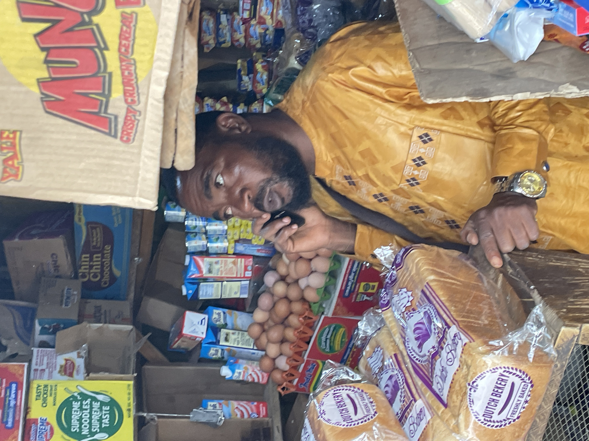 The image shows a man with dark skin and a full beard, wearing a mustard-yellow shirt with geometric patterns near the shoulders, is seen talking on a phone. He is in a small shop or store in Jos, Nigeria. He has a gold wristwatch on his left wrist and is surrounded by various items for sale, including bread, eggs, and packaged foods. A box of "MUN" cereal and "Supreme Noodles" are visible, along with "Chin Chin Chocolate" snacks. The bread appears to be of the "Dutch Bekery" brand. The scene is somewhat cramped, with shelves and stacked boxes filled with items.