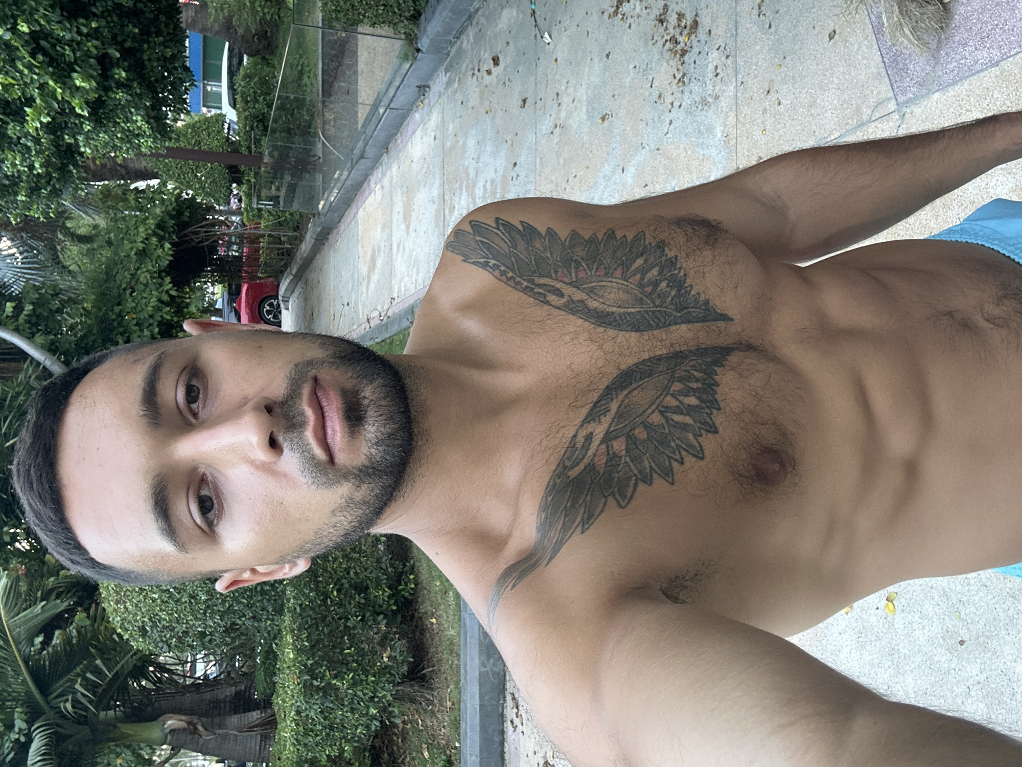 A shirtless man with a beard and a wing tattoo on his chest stands in a park. He is facing the camera and smiling slightly. The park has palm trees, manicured bushes, and a paved walkway. In the background, there is a red car and other vehicles. The image appears to have been taken in Nha Trang Ward, Vietnam.