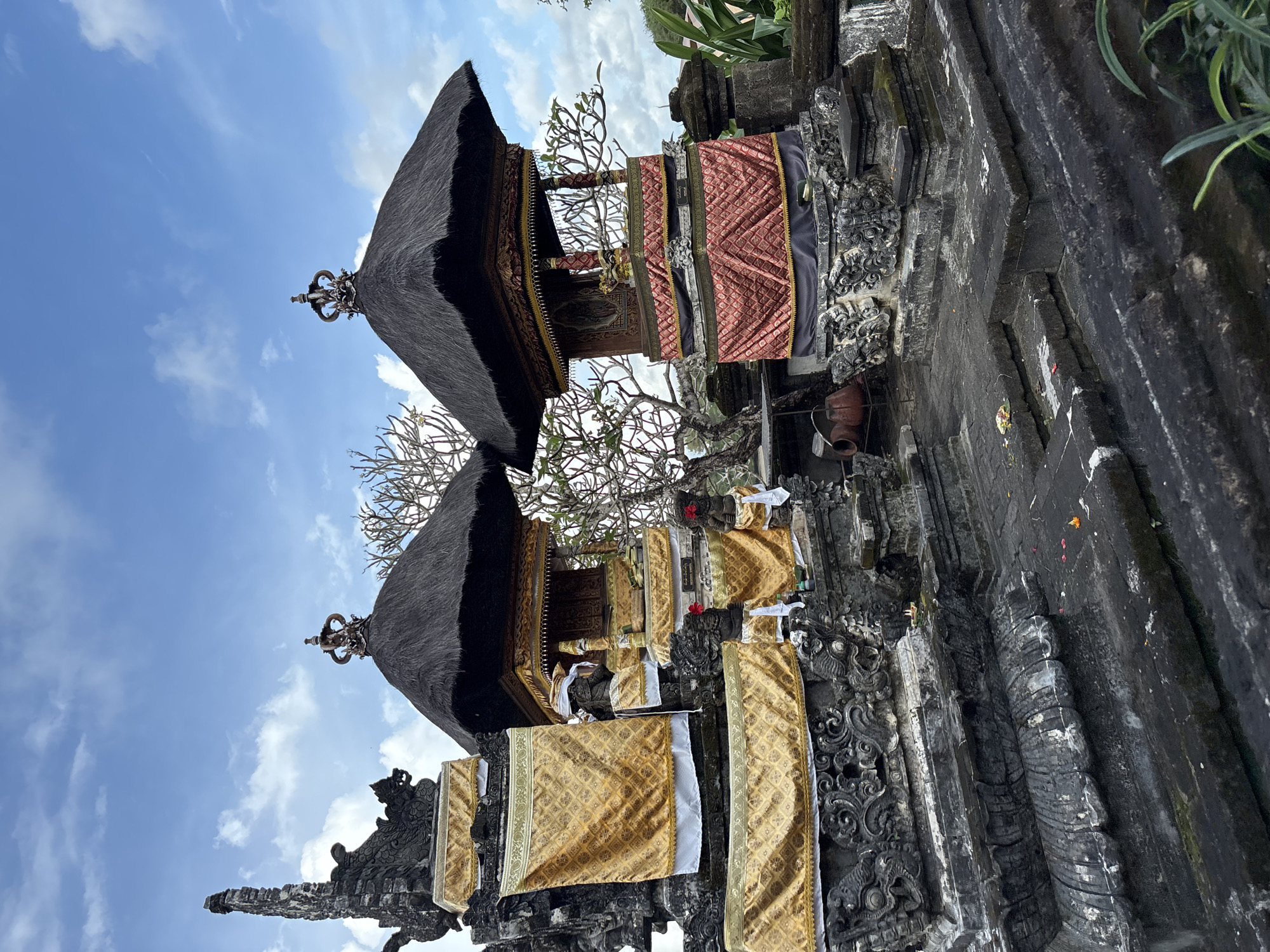 This image depicts Balinese shrines in Kuta Selatan, Indonesia. The shrines are of dark stone with intricate carvings and feature thatched roofs topped with metal ornaments. Decorative fabrics of gold and red adorn the structures. The sky is visible in the background, with scattered clouds. A flowering tree is nestled between the shrines.