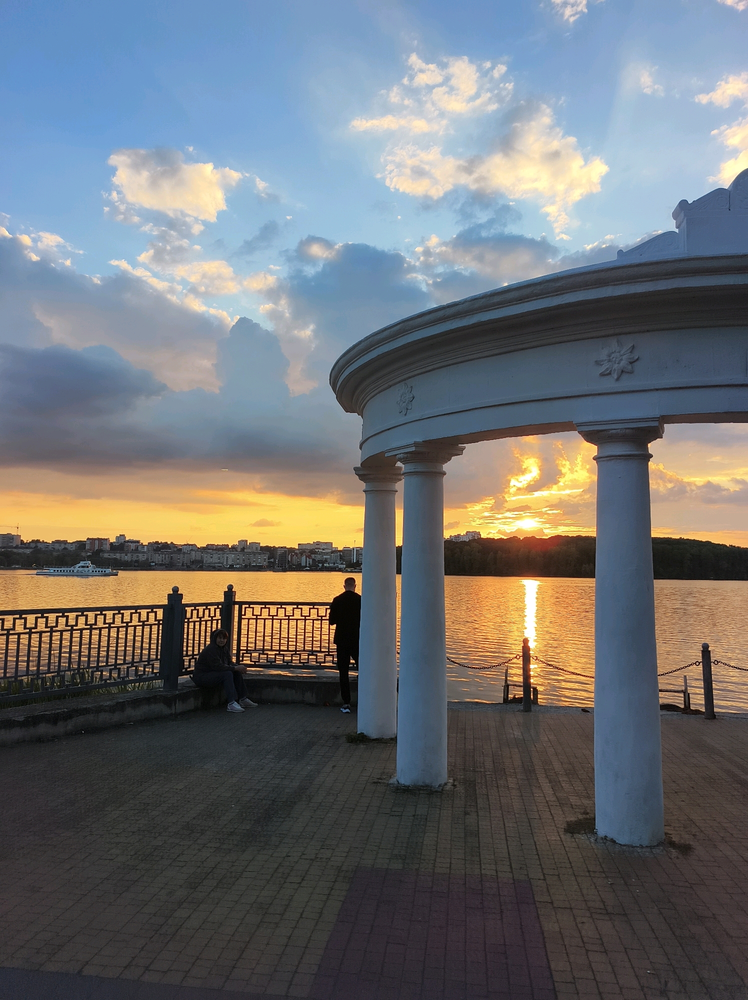 The image depicts a sunset over the Ternopil Pond in Ternopil, Ukraine. In the foreground, a white rotunda with several columns stands on a paved walkway. Two people are visible on a bench to the left of the walkway, one sitting and the other standing. A boat is sailing on the pond in the middle ground, with a city skyline in the background. The sky is filled with dramatic clouds, illuminated by the setting sun, casting a warm glow on the water. The overall scene conveys a sense of tranquility and natural beauty.