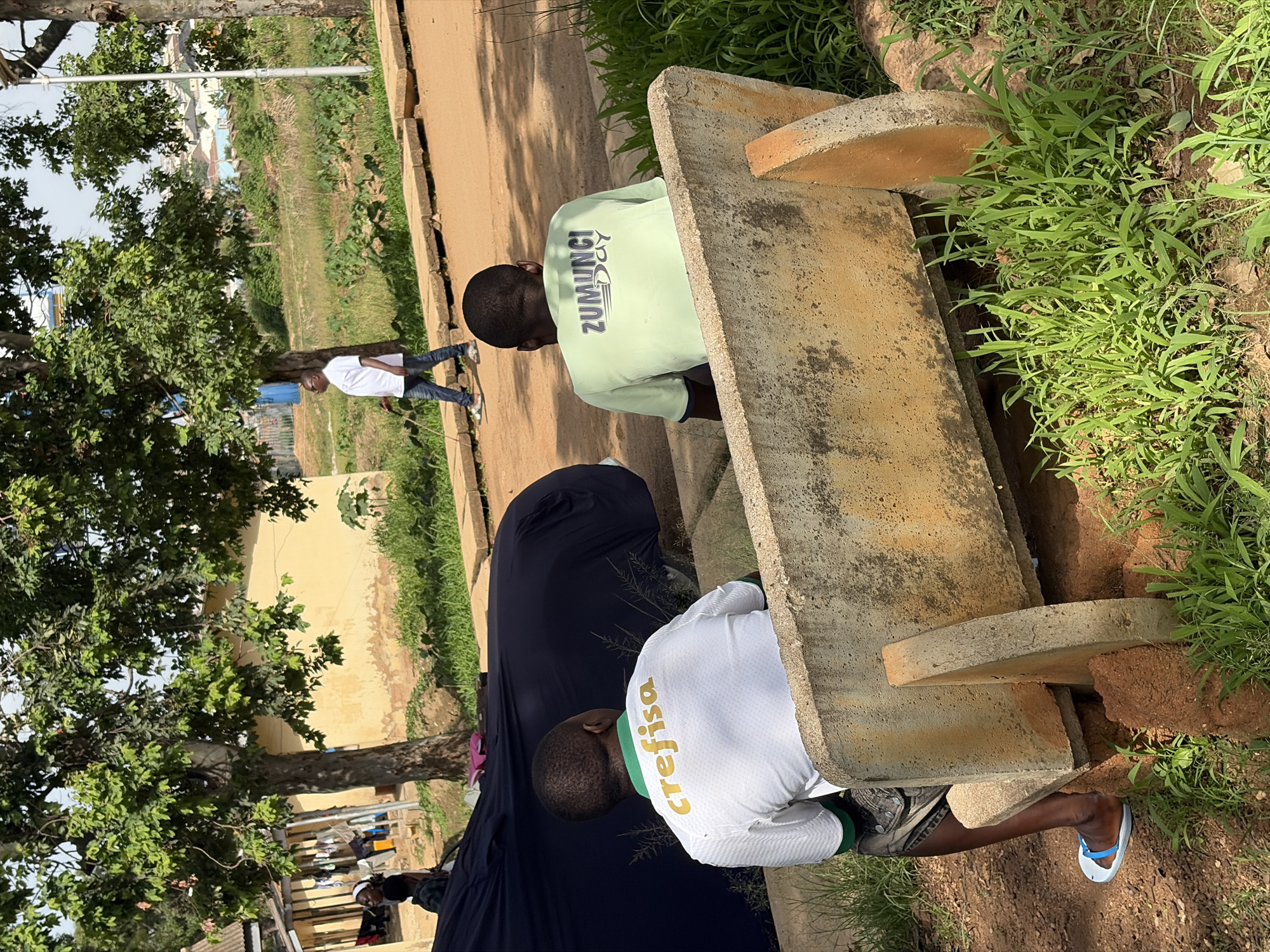 This is a medium shot, eye-level, outdoor photograph taken in the city of Rafin Par, Nigeria. In the foreground, two men are seated on a concrete bench, viewed from behind. The man on the left wears a white and green t-shirt with gold lettering that reads "crefisa". The man on the right wears a light green t-shirt with black lettering that reads "ZUMUNCI DAY". Both men are wearing shorts. The man on the left wears light blue and white flip-flops. The bench is weathered and stained with orange dirt. Behind the bench, to the left, is a dark blue car cover over an object, likely a car. Further in the background, there are trees with green foliage and a building with a pale yellow exterior. A dirt path and concrete walkway are visible. In the distance, more buildings and vegetation can be seen, with a hazy sky above. To the right of the bench, green grass and some dry vegetation are present. In the mid-ground, a man in a white t-shirt and blue jeans walks along the concrete path, holding a walking stick. He wears glasses.