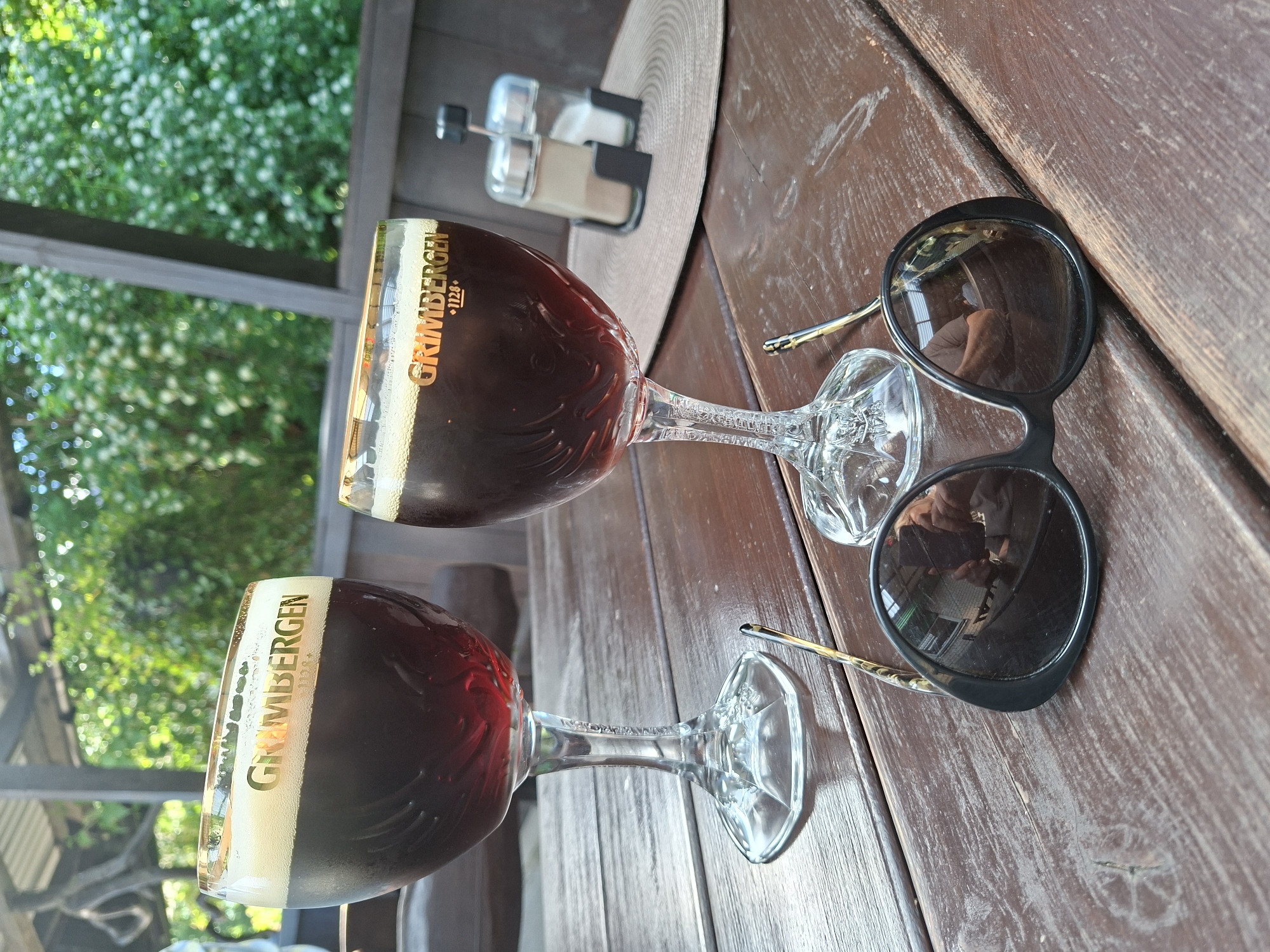Two glasses of dark beer with foam tops sit on a rustic wooden table. The glasses have gold rims with the Grimbergen logo printed on them. In the foreground, a pair of black sunglasses rests on the table, reflecting a person taking the photograph. In the background, a blurred view of greenery and outdoor seating suggests a beer garden or outdoor dining area. The scene is set outdoors, possibly at a restaurant or cafe in Mikytai, Lithuania, given the context.