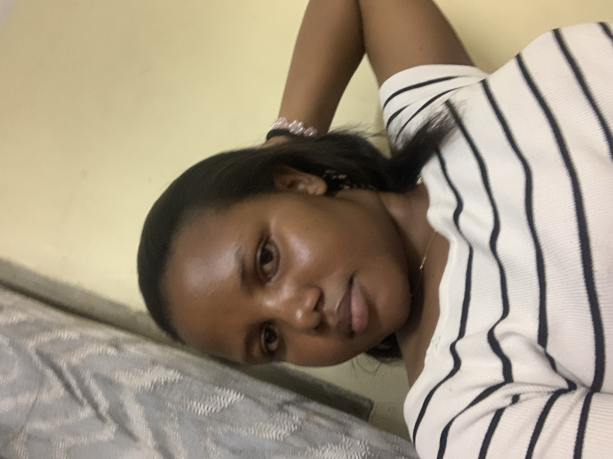 A woman is seen indoors lying on a bed with her arm raised, wearing a striped white and black shirt. The background features a cream-colored wall and a textured bed cover. The location is unspecified but hypothesized to be in the city of Dar es-Salaam, Tanzania.