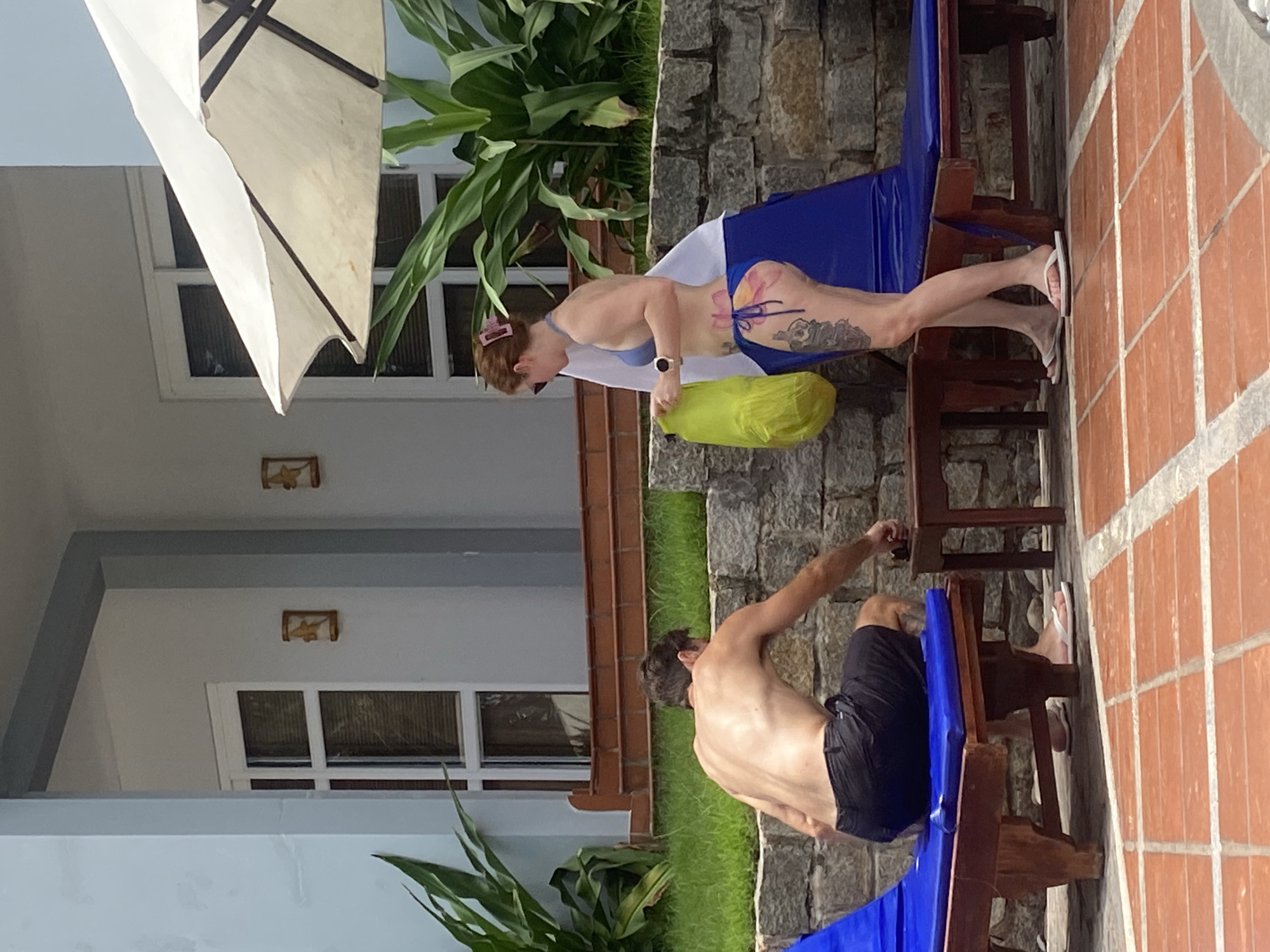 This image depicts an outdoor scene at what appears to be a resort or hotel in Phu Thuy Ward, Vietnam, under bright daylight conditions.
In the foreground and center, a woman with auburn hair is standing by a blue lounge chair, viewed from her left side. She is wearing a light blue bikini top and a blue bikini bottom. Distinctive tattoos are visible on her left leg and hip, including a large pink floral design on her hip/buttock and intricate patterns on her thigh. She has sunglasses on her head, a watch on her left wrist, and white flip-flops on her feet. A bright yellow bag rests on the lounge chair beside her, and she seems to be reaching for or adjusting something.
To the left and slightly behind the woman, a shirtless man is seated on another blue lounge chair, facing away from the camera. He is wearing dark swim trunks and white flip-flops. His posture suggests relaxation, looking towards a stone wall or the ground.
The immediate setting features a patterned tiled ground in reddish-brown hues where the lounge chairs are placed. Behind the subjects, a low stone wall transitions into a strip of green grass and lush tropical plants. The background consists of a multi-story building with light-colored walls (white or light blue) featuring white-framed windows and a balcony structure. A large, partially visible white sun umbrella is positioned in the upper left. Small decorative wall lights are also visible on the building's facade. The scene conveys a relaxed, leisurely atmosphere typical of a vacation spot. No discernible text is visible.