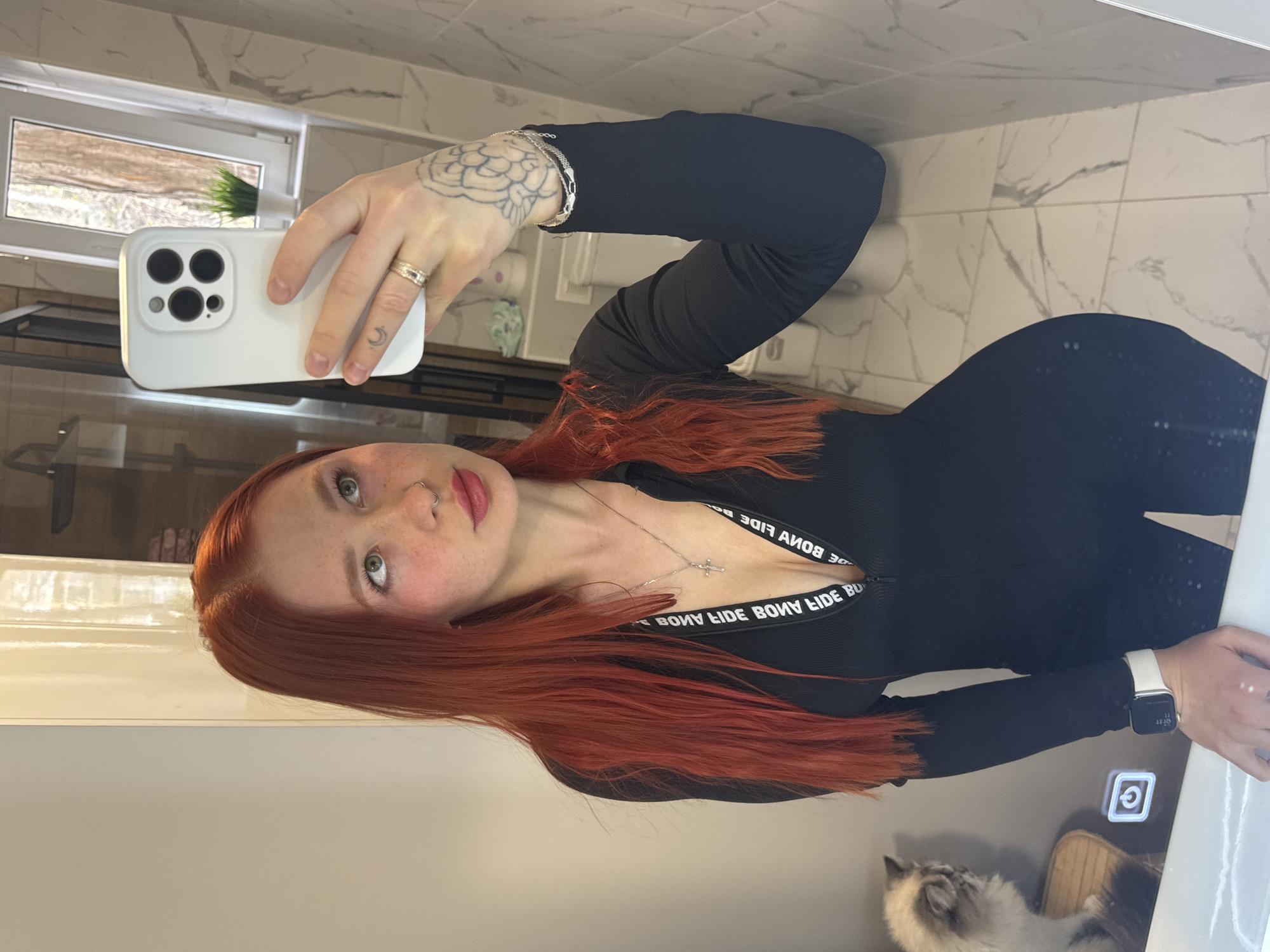 A young woman with long, red hair is taking a selfie in a bathroom. She is wearing a black, long-sleeved outfit with white lettering that reads "BONA FIDE" repeatedly around the chest. She is holding a white smartphone in her left hand, showing a multi-camera setup. She has light skin, freckles, and wears lipstick, and has a nose ring. She is wearing a ring on her left hand and a smartwatch on her right wrist.
The bathroom features white tiled walls and a counter with an integrated sink. A light is reflected on the sink surface. A cat is partially visible at the bottom of the image. There is a window above a tiled backsplash visible in the background. The location cannot be determined from this image.