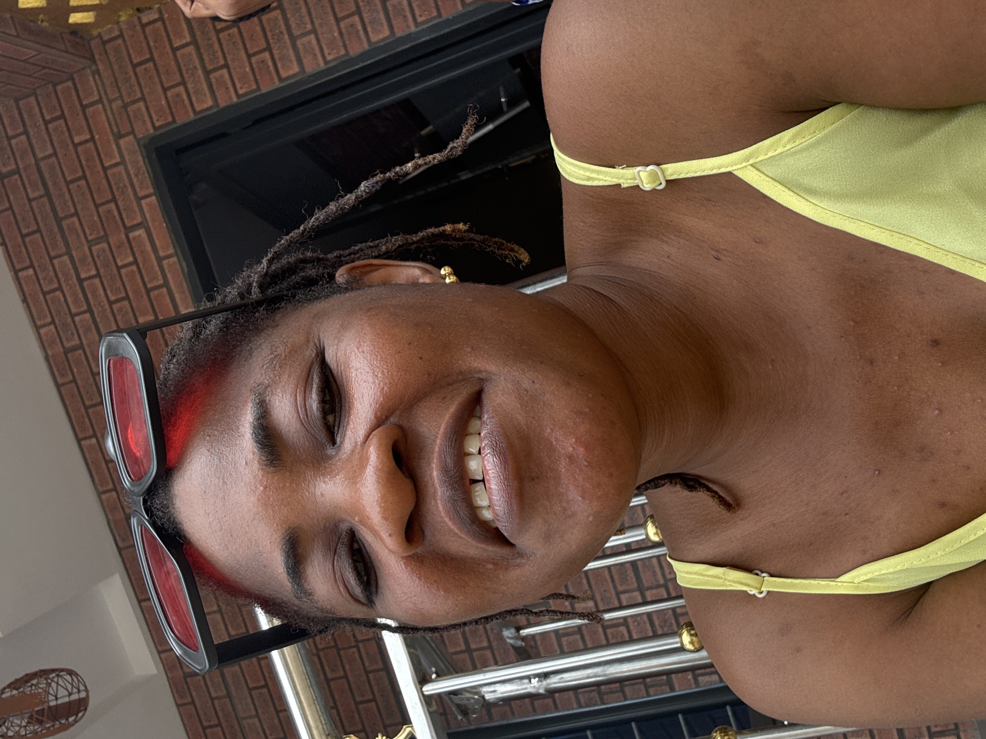 A young Black woman is shown in a close-up, selfie-style shot. She is outside, with a brick building and a metal railing visible in the background. The woman is wearing a light yellow top with thin straps and red-rimmed sunglasses pushed up on her forehead. Her hair is styled in dreadlocks, with some strands framing her face. She has a gold earring in her visible ear and her teeth are slightly parted as she smiles. The overall impression is casual and relaxed, suggesting the photo was taken during a pleasant day outdoors. This scene is located in Ajara, Nigeria.