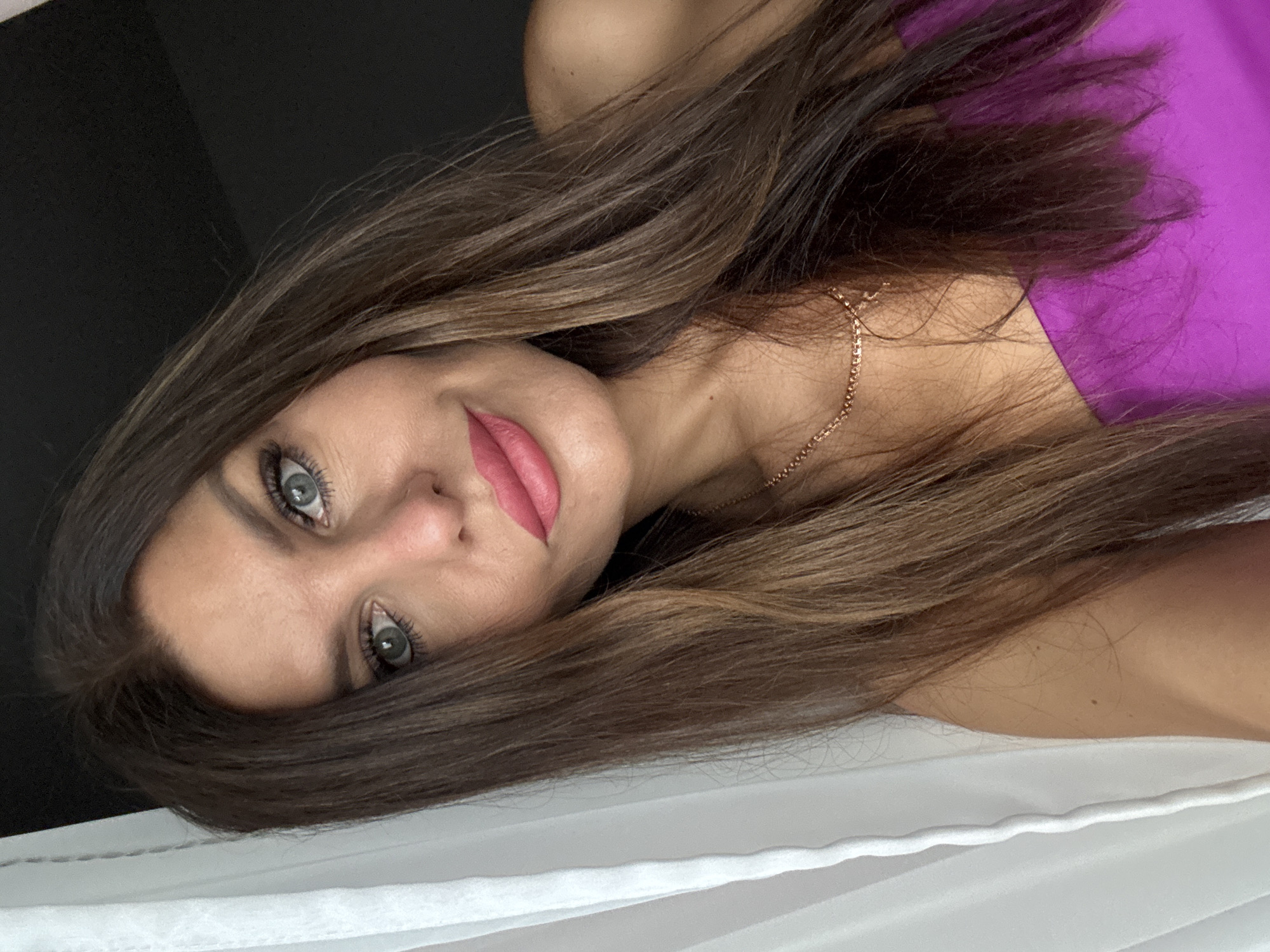 A woman with long, dark brown hair, blue eyes, and pink lipstick poses for a selfie indoors, leaning against a white curtain. She is wearing a strapless purple top and a gold necklace. The background is dark.