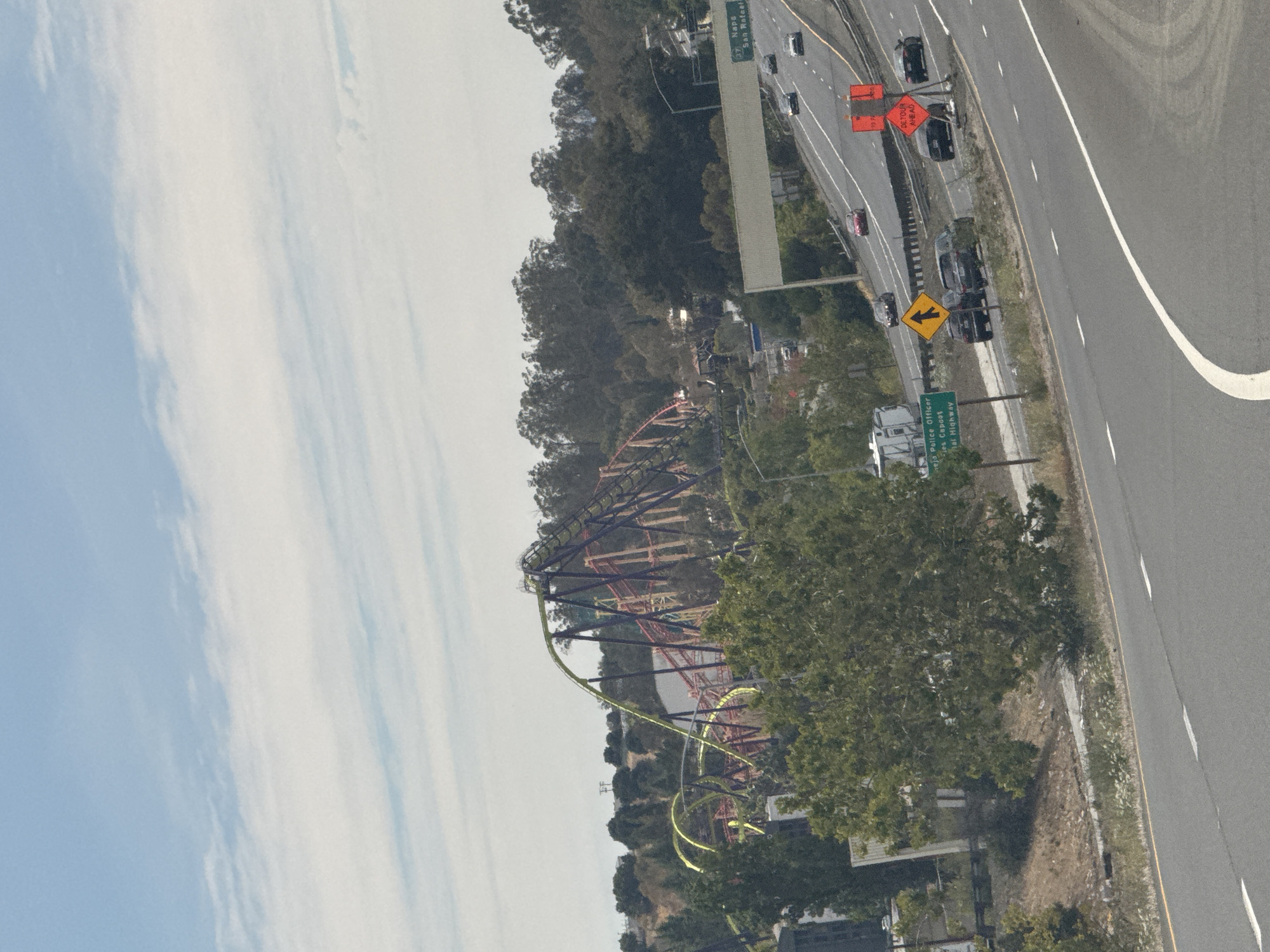 The image captures a view of the roller coaster at Six Flags Discovery Kingdom in Vallejo, California, USA, from a highway. The roller coaster, with its yellow, purple, and red tracks, is prominent in the background, set against a backdrop of green trees and a cloudy sky. In the foreground, a multi-lane highway is visible with several cars driving on it. Road signs are present, including one indicating "Napa San Rafael" and another for "Detour Ahead." A green sign for "Vallejo Police Officer" is also visible. The overall scene depicts a sunny day with a mix of amusement park and highway elements.