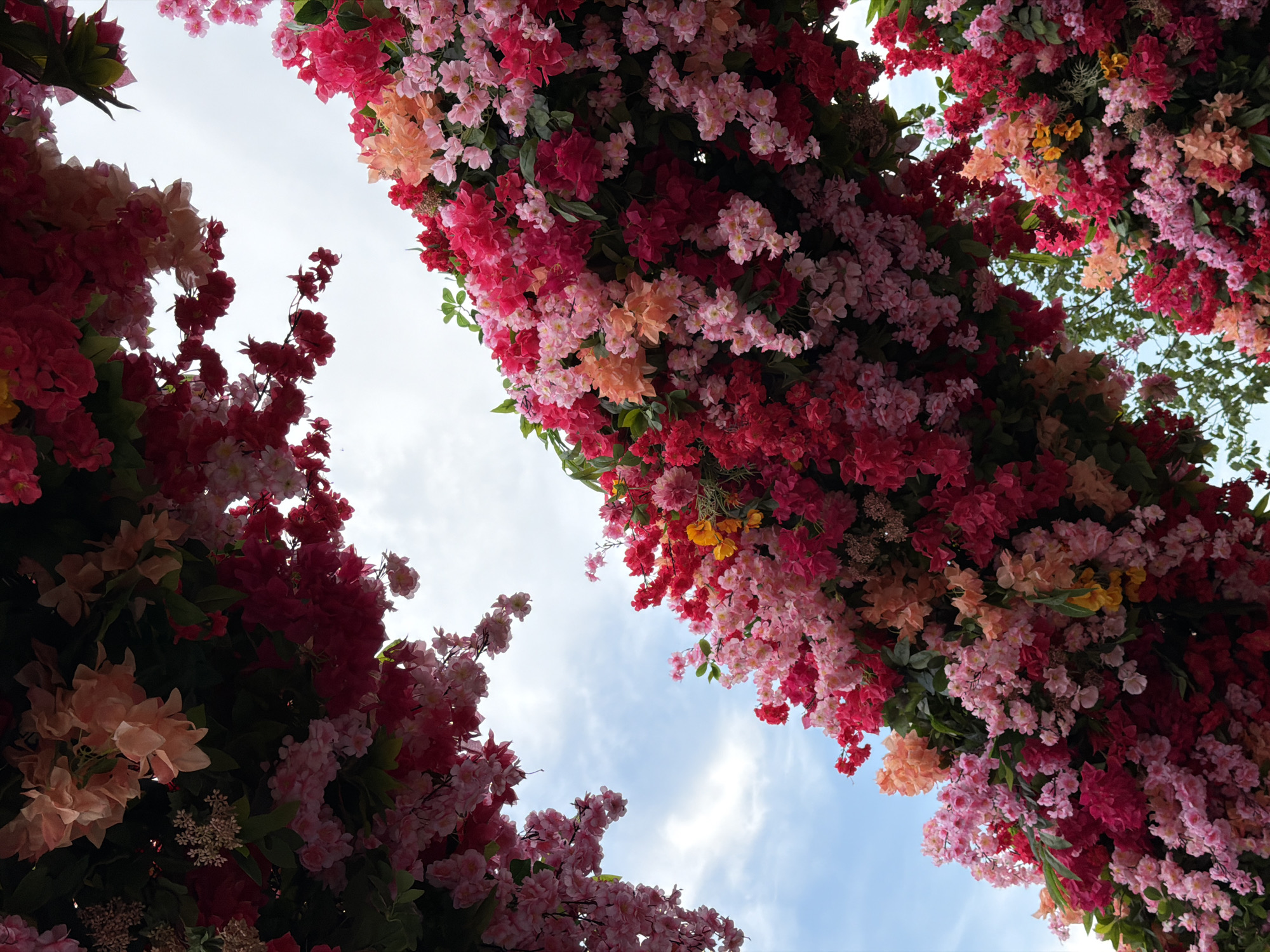 A low-angle shot looks up at a densely flowered trellis set against a partly cloudy sky. The trellis is a mix of pink, red, peach, and white flowers, appearing to be a constructed floral display. The sky is light blue with patches of white clouds visible between the flowers. The scene has no people or animals. Based on similar displays and the prevalence of such decorations in urban areas, this image could be from a park or public space in Moscow, Russia, known for its elaborate floral displays.
