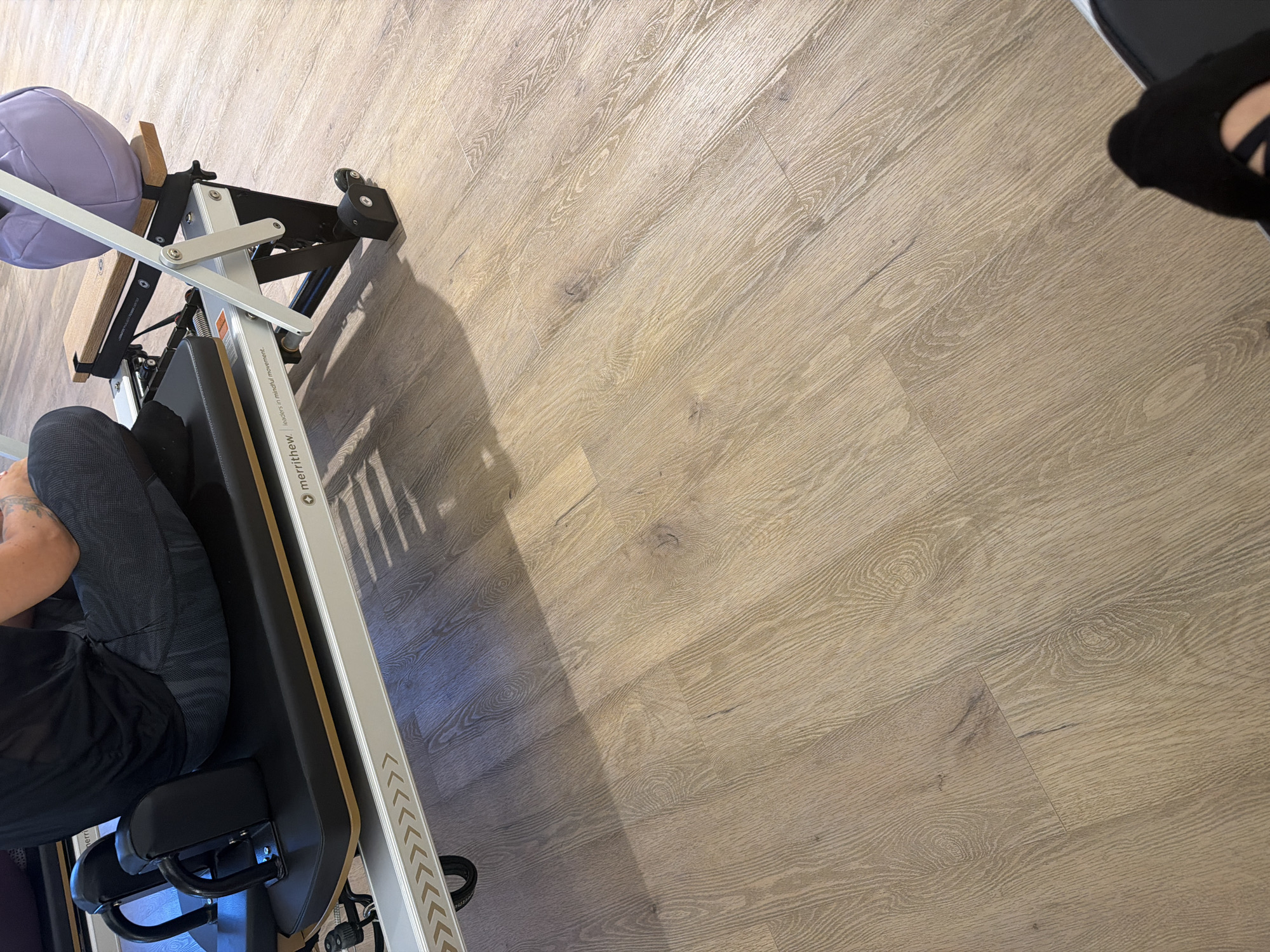 The image shows a person sitting on a piece of gym equipment that appears to be a Pilates reformer. The person is wearing a black t-shirt and dark patterned leggings. The equipment is black with some metallic accents and a wooden seat. A light purple exercise ball is resting on the equipment behind the person. The scene is indoors, with a light-colored wooden floor. The lower right corner of the image shows a black sock. The image does not contain any information that can identify its location within Boreti, Montenegro.