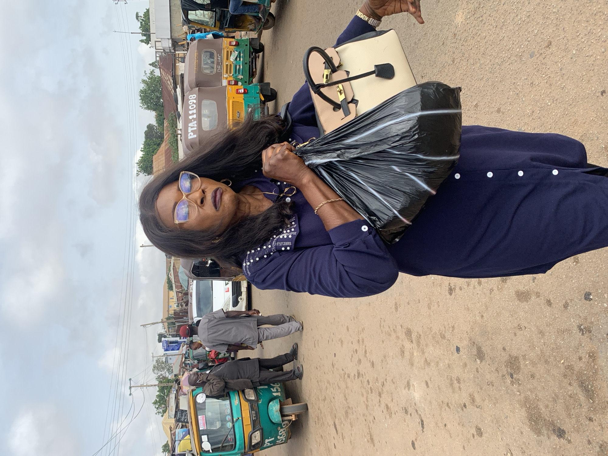 This is a full-body, eye-level, outdoor shot of a woman with dark skin, black hair, and glasses walking on a dirt road. The woman is wearing a navy blue long-sleeved dress with white buttons down the front. She is carrying a black plastic bag filled with items in her right hand and a light-colored handbag with black trim and gold hardware in her left arm. The woman's head is turned slightly to her right, and she is looking forward with a serious expression. The background of the image shows a busy street scene, with several three-wheeled vehicles, commonly known as "Keke" or "Maruwa," and other vehicles. There are also people in the background, some appearing to be vendors or passengers. The environment appears to be a market or transportation hub in Gura Top, Nigeria, characterized by a dusty road, various structures, and utility poles with wires overhead. The sky is overcast, suggesting a cloudy day.