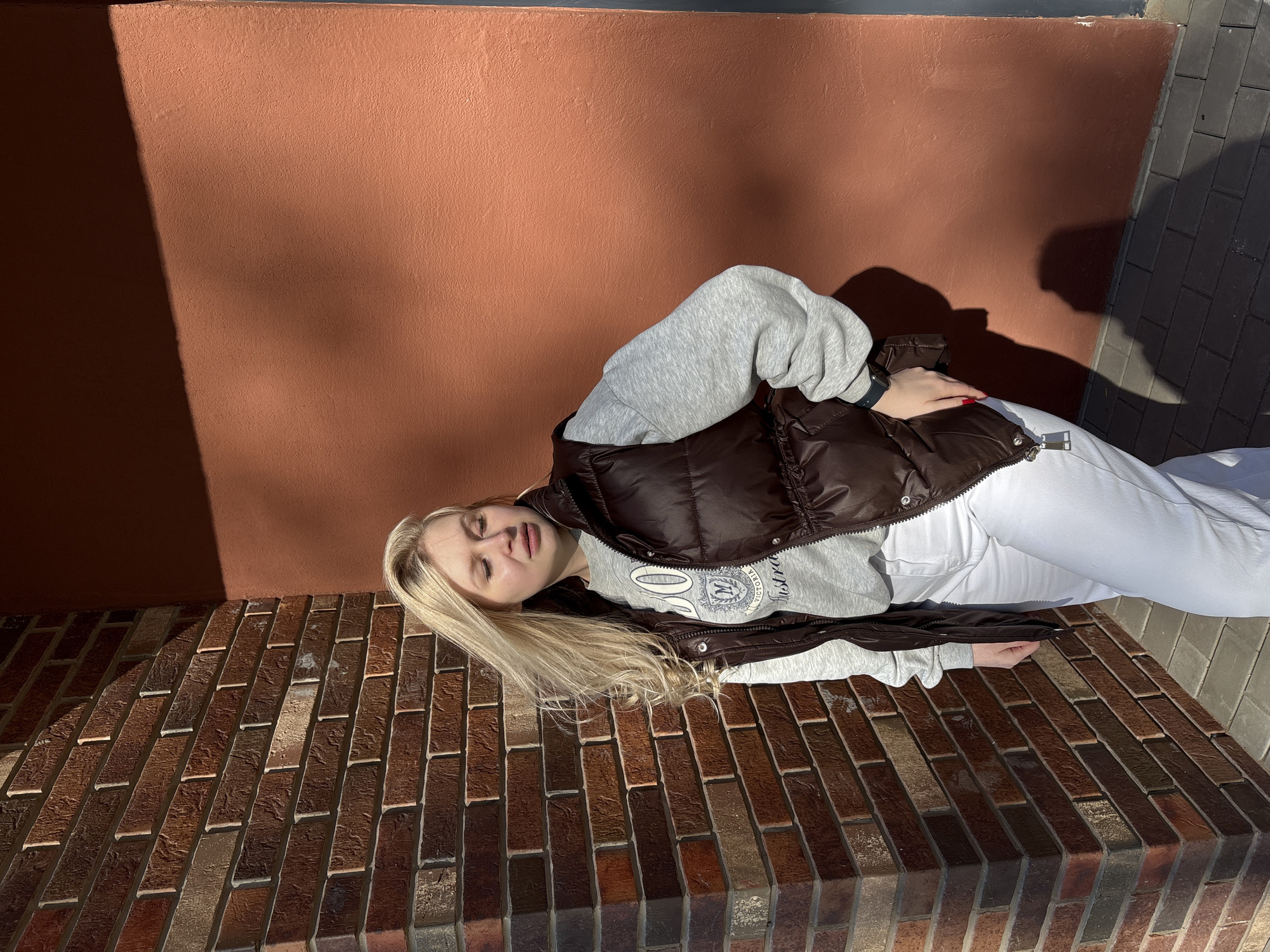 A blonde woman lies on her side on a brick ledge in front of a terracotta-colored wall with dark shadows. She is wearing a gray sweatshirt with a logo, a dark brown puffer vest, and white pants. The brick ledge extends to the right, where it meets a gray concrete surface. The image is rotated clockwise. There is no evidence in the image to confirm the location is Izhevsk, Russia.
