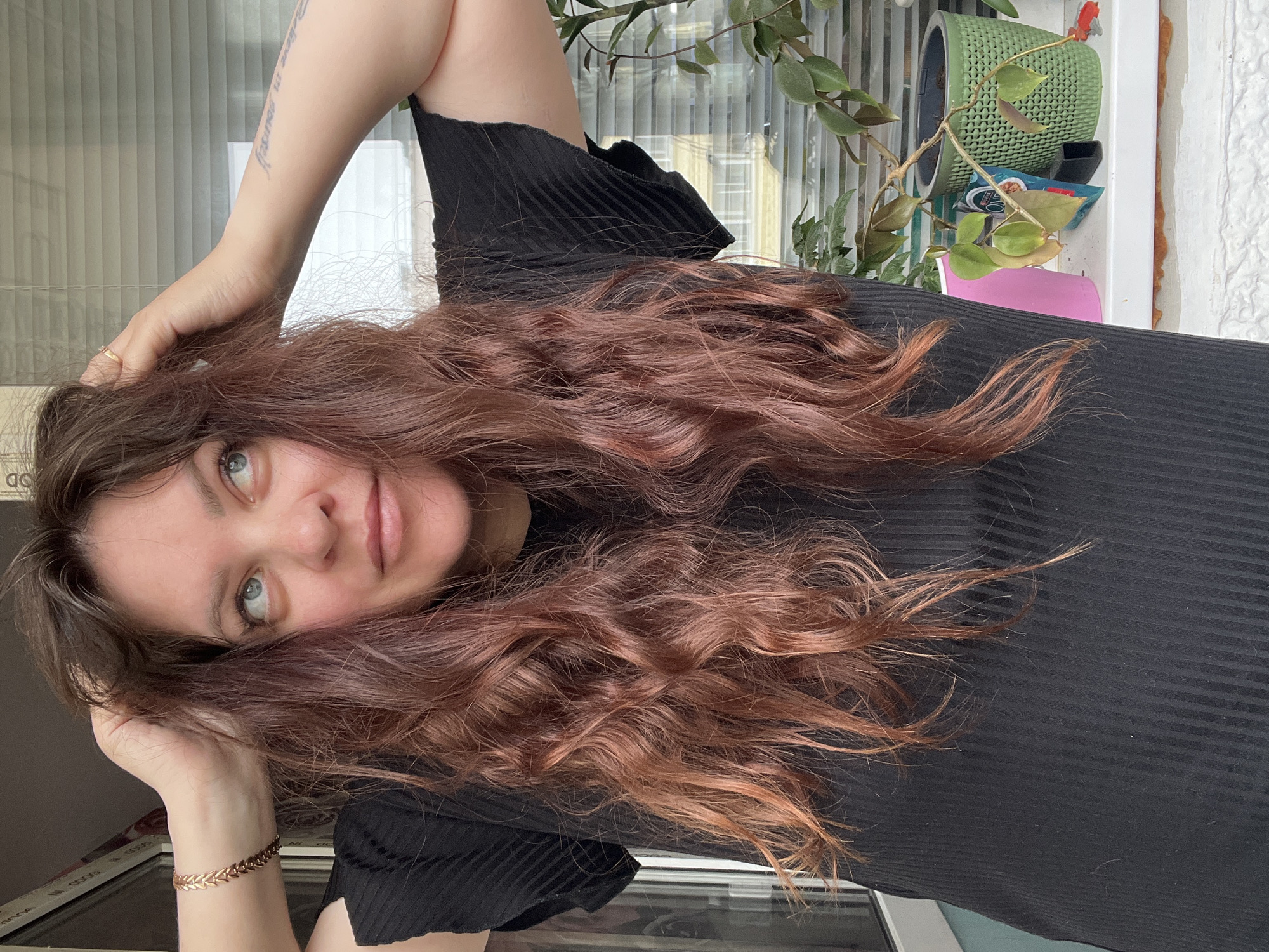 A close-up portrait features a young woman with long, voluminous, wavy reddish-brown hair, positioned as if reclining. Her blue-green eyes are directed upwards and to the right, accompanied by a soft, gentle smile. She is wearing a black ribbed top. One hand is near her head, lightly touching her hair, with a subtle gold-colored ring visible on her left ring finger. Her other arm is bent, revealing a rose-gold bracelet with a leaf-like pattern on her right wrist and a tattoo on her inner bicep that reads "Believe in yourself."
The setting is indoors, with natural light suggesting it is daytime. Behind her, a window with vertical blinds offers a glimpse of a building exterior. An indoor plant with green leaves, a green textured planter, and a pink object are visible in the background, implying a domestic or personal space in Noviy, Russia. The woman appears calm, relaxed, and thoughtful.