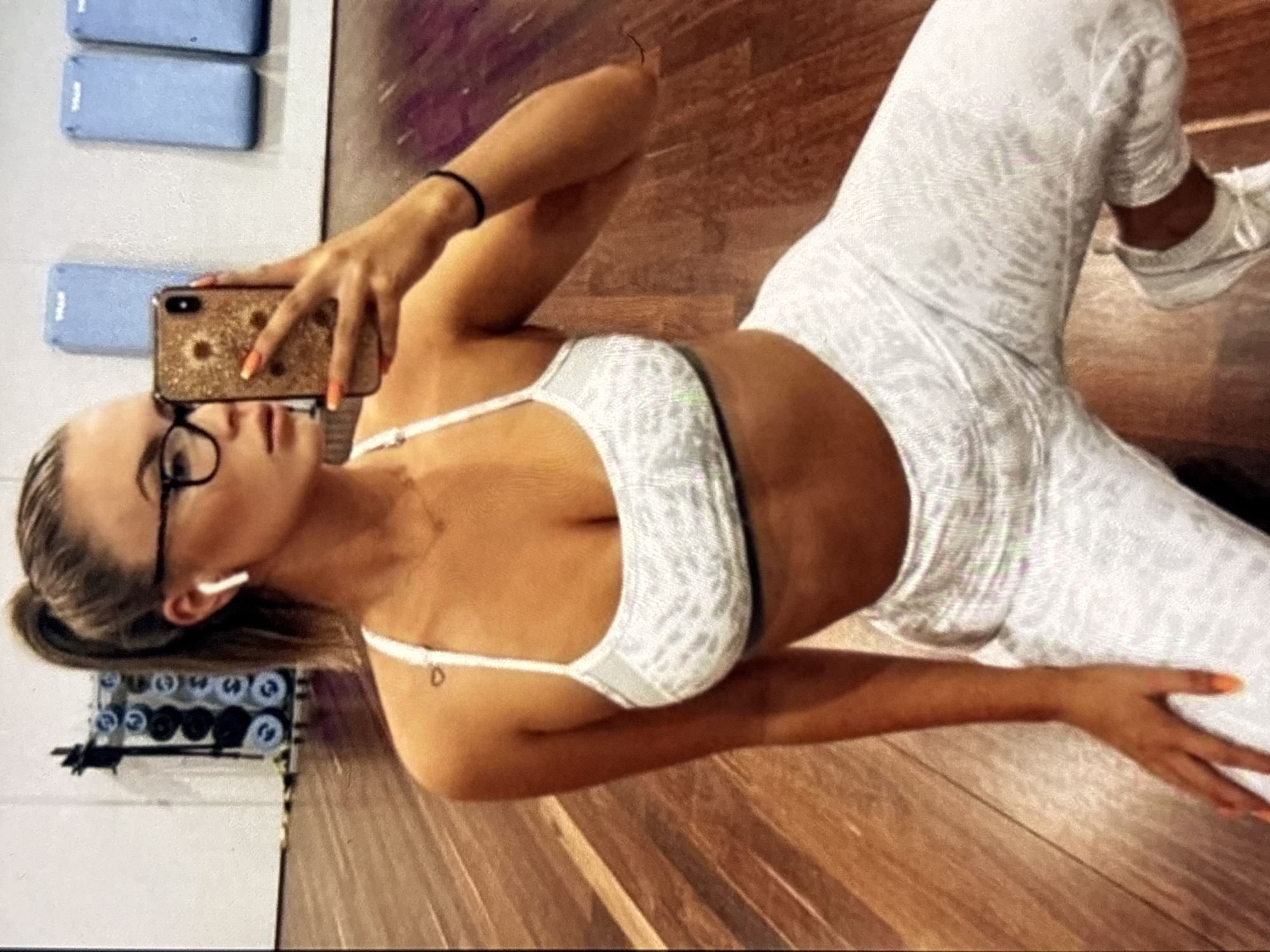 A woman with a high ponytail and glasses is kneeling on a wooden gym floor, taking a selfie with her smartphone. She is wearing a white sports bra and white leggings with a light grey leopard-like print. She has on white sneakers and orange nail polish. The gym has weight racks and blue wall-mounted mats in the background. She has an earbud in her left ear and a bracelet on her right wrist.