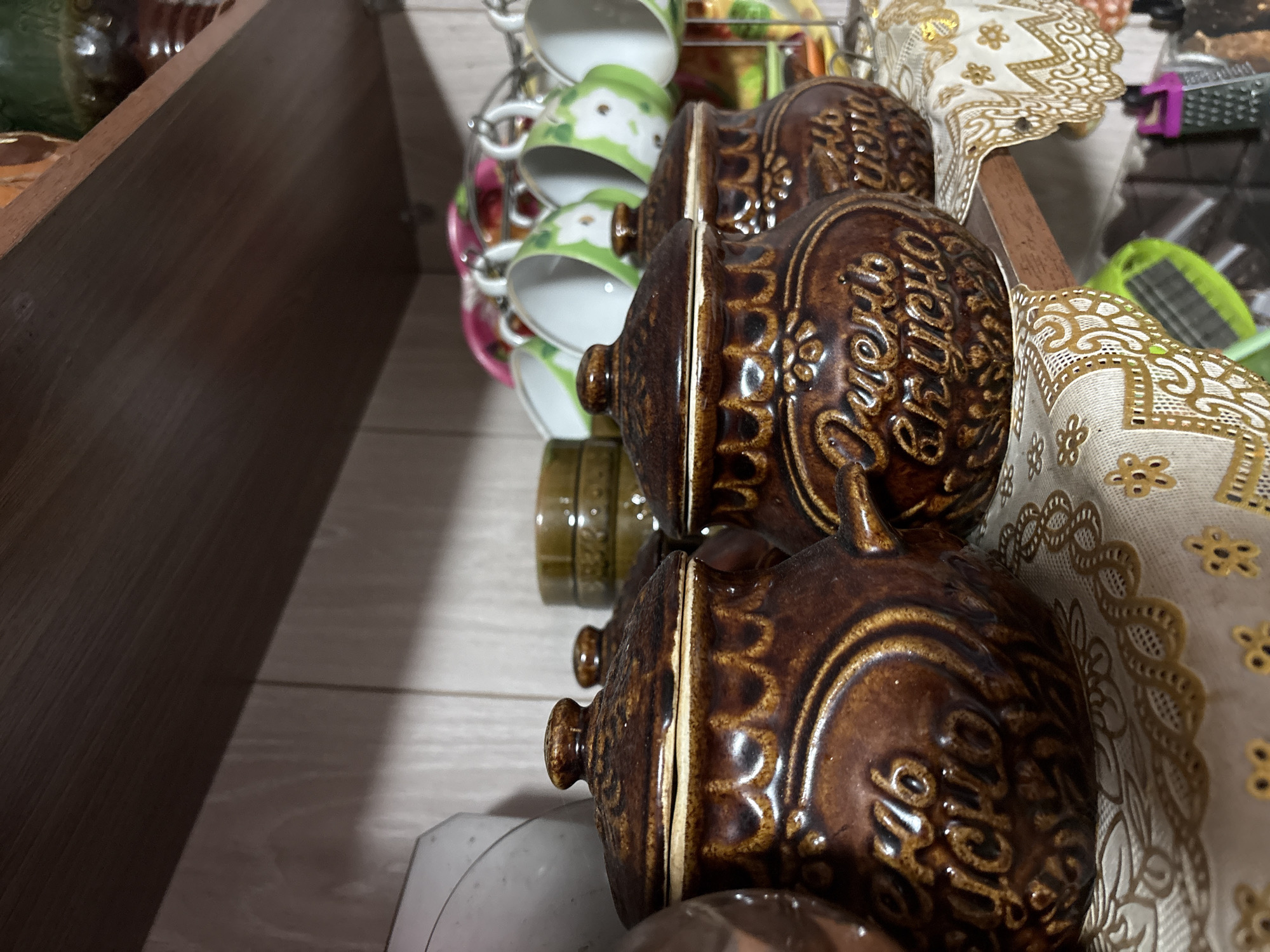 The image shows a close-up view of three ceramic pots with lids on a wooden shelf. The pots are dark brown and have ornate relief patterns. The pot in the foreground on the left has Cyrillic writing in yellow on it, which appears to read "Хлеб всему голова" (Bread is the head of everything). The pot in the middle has similar Cyrillic writing, possibly "Очень вкусно" (Very tasty). In the background, there is a rack of colorful mugs, some green with white floral patterns, and others with pink floral patterns. The shelf is covered with a decorative cloth with gold trim. The overall impression is that this is a domestic setting, likely a kitchen or pantry, in the city of Vetluzhsky, Russia.