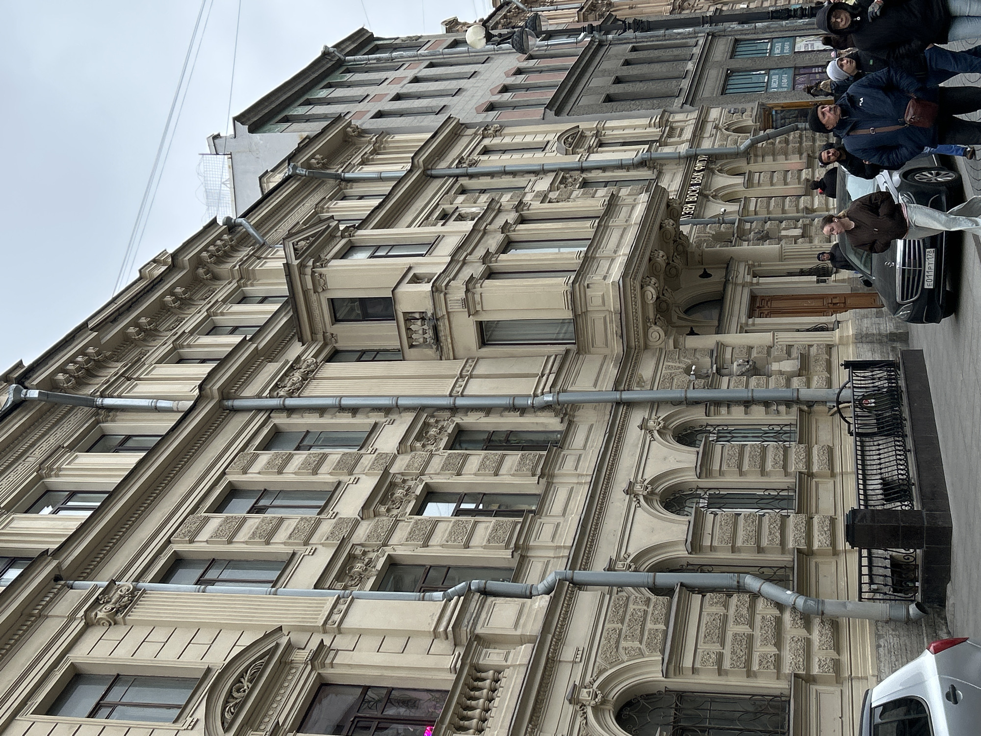 An outdoor, slightly tilted shot captures a street scene dominated by a large, ornate multi-story building under an overcast sky, suggesting cool weather. The building features elaborate classical or neoclassical architectural details, including decorative cornices, pilasters, patterned stonework, and numerous windows. Several grey drainpipes run horizontally and vertically across the facade.
On the sidewalk below, a group of people is visible, mostly dressed in jackets and hats, indicating cooler temperatures. Some are standing, while others appear to be walking. A man in a dark blue jacket and hat with a brown shoulder bag looks towards the right. A woman in a brown top and light pants is partially visible in the foreground, seemingly looking at her phone.
Parked on the street is a black Mercedes-Benz sedan, with its license plate "E011PT 178" clearly visible. The number "178" is a regional code for Saint Petersburg, Russia. Part of a white vehicle is also visible in the bottom right corner.
Visible text on the building includes a sign above an entrance that reads "МУЗЕЙ ВОСКО ВЫХ ФИГУР" (MUSEUM OF WAX FIGURES). Higher up on the building, a partial sign for "NEW YORK STORE" can be seen. Overhead wires stretch across the sky above the building's roofline. The overall scene depicts a typical urban environment in Saint Petersburg, characterized by historic architecture and street activity.